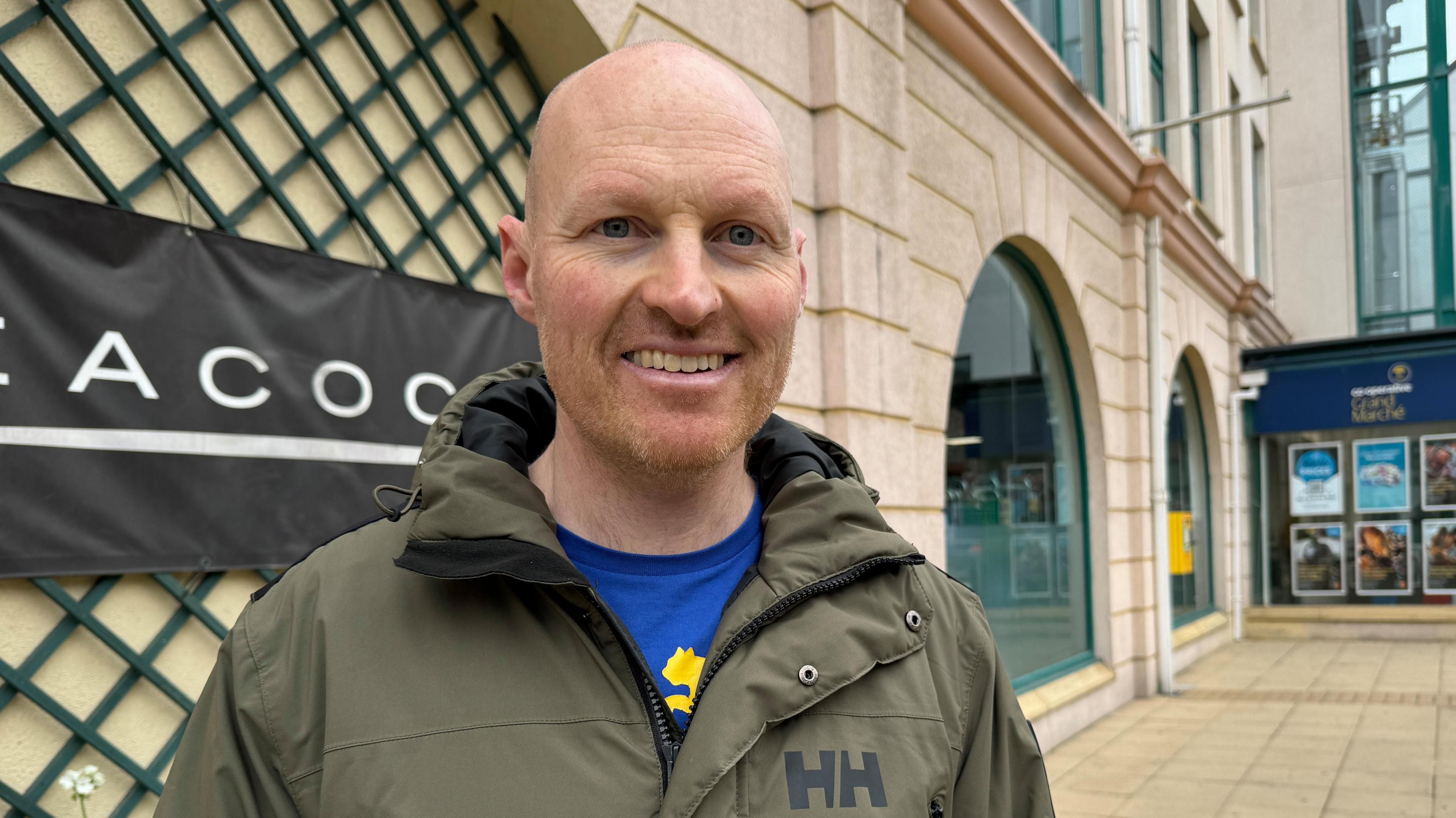 Man in green coat stands outside a shopping centre.