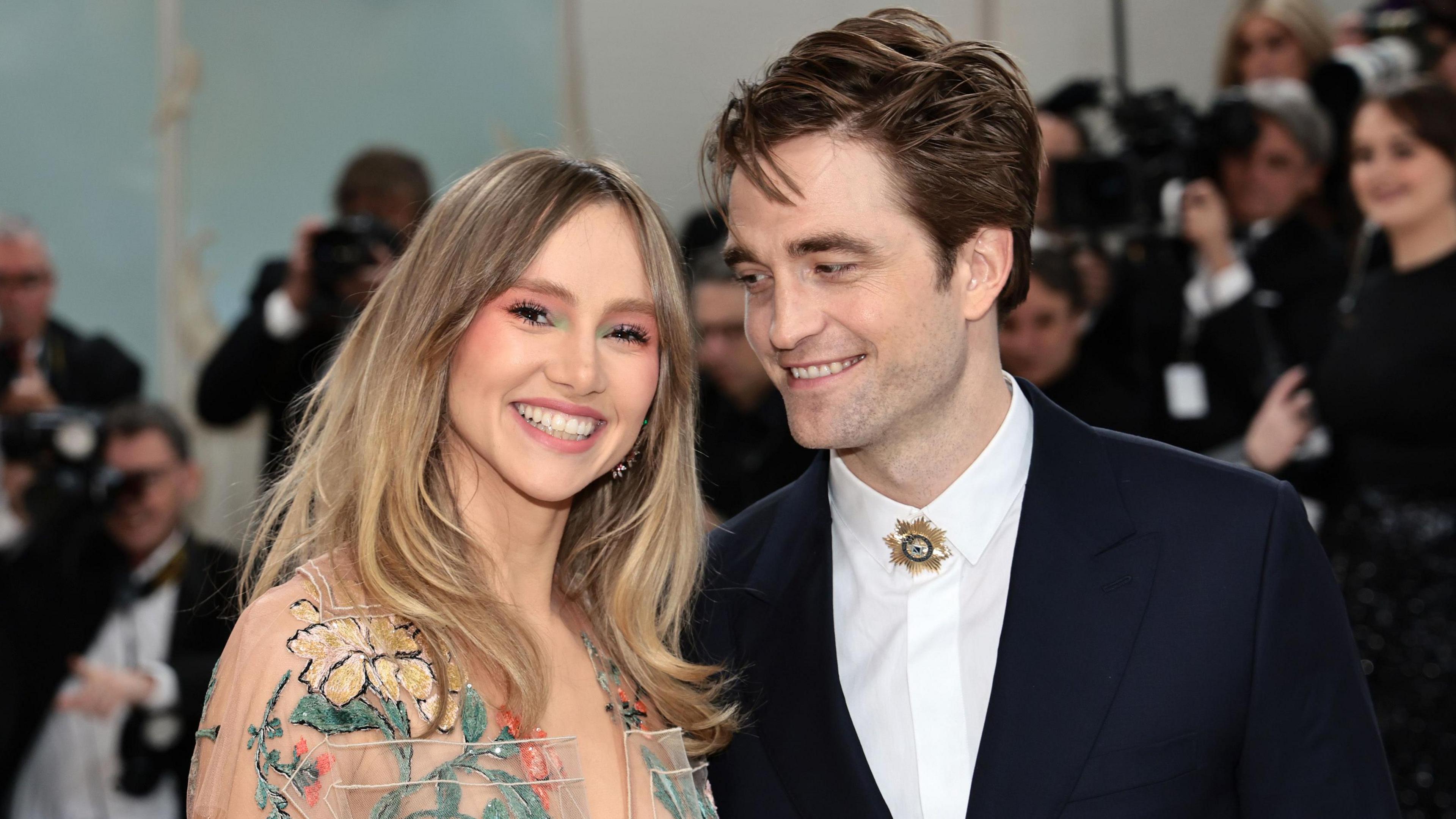 Suki Waterhouse and Robert Pattinson on the red carpet for the 2023 Met Gala. Suki and Robert are both in their 30s and dressed up for the event - Suki in a sheer floral dress and Robert in a white shirt and dark suit with a broach replacing his tie. He smiles at Suki, who is smiling at the camera. Photographers can be seen behind them in the background 