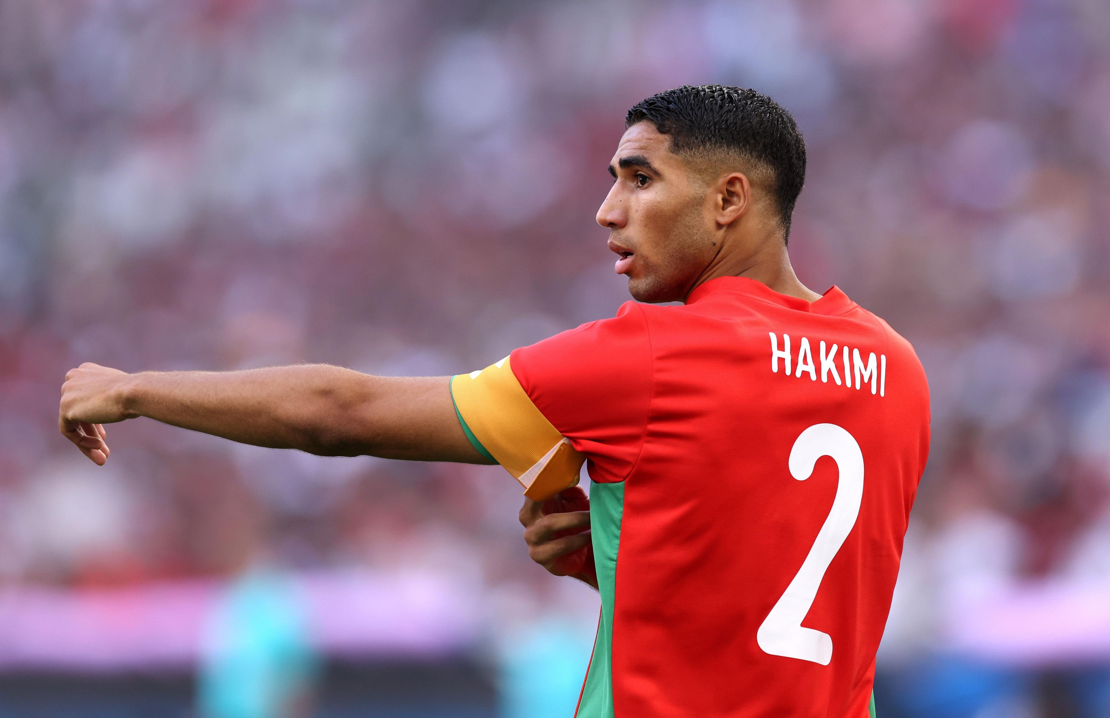 Achraf Hakimi shown from behind and from waist up wearing a red Morocco jersey with green stripes under the arm and a white number two on the back. He is pictured turning sideways towards the camera during a match and pulling an orange captain's armband up on his left arm