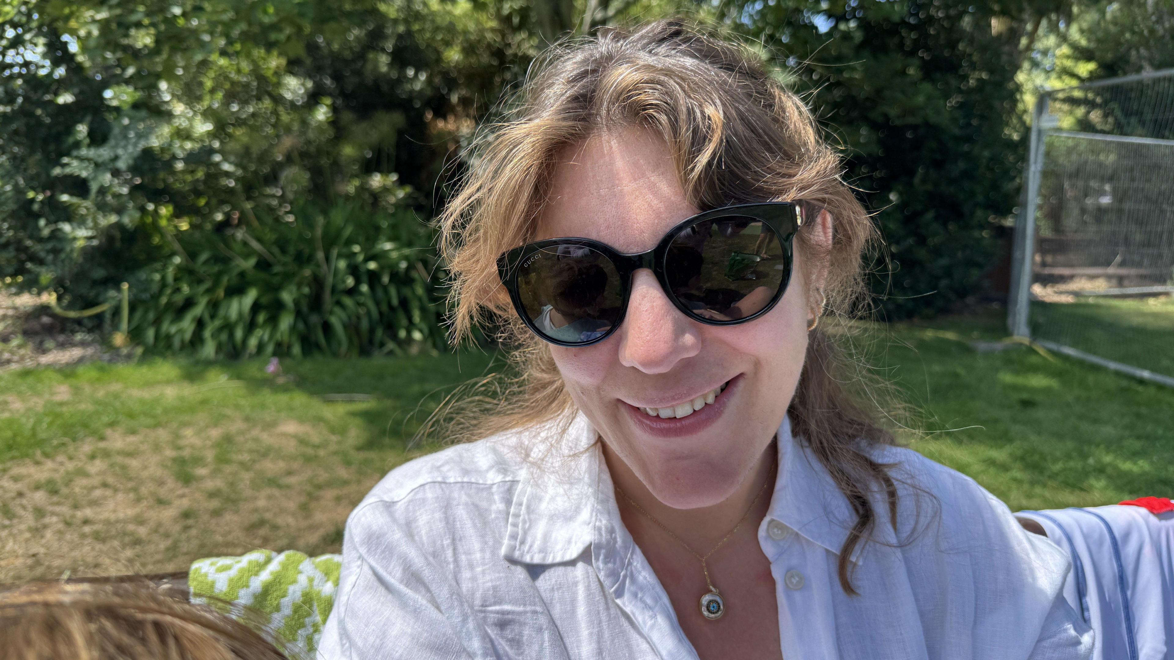 A woman sits in a park surrounded by trees and bushes. She has light brown hair, loosely tied back and she is wearing sunglasses. She is wearing a crisp white button-up shirt paired with a circular pendant necklace. A green and white blanket is draped over the back of the bench she is sitting on.