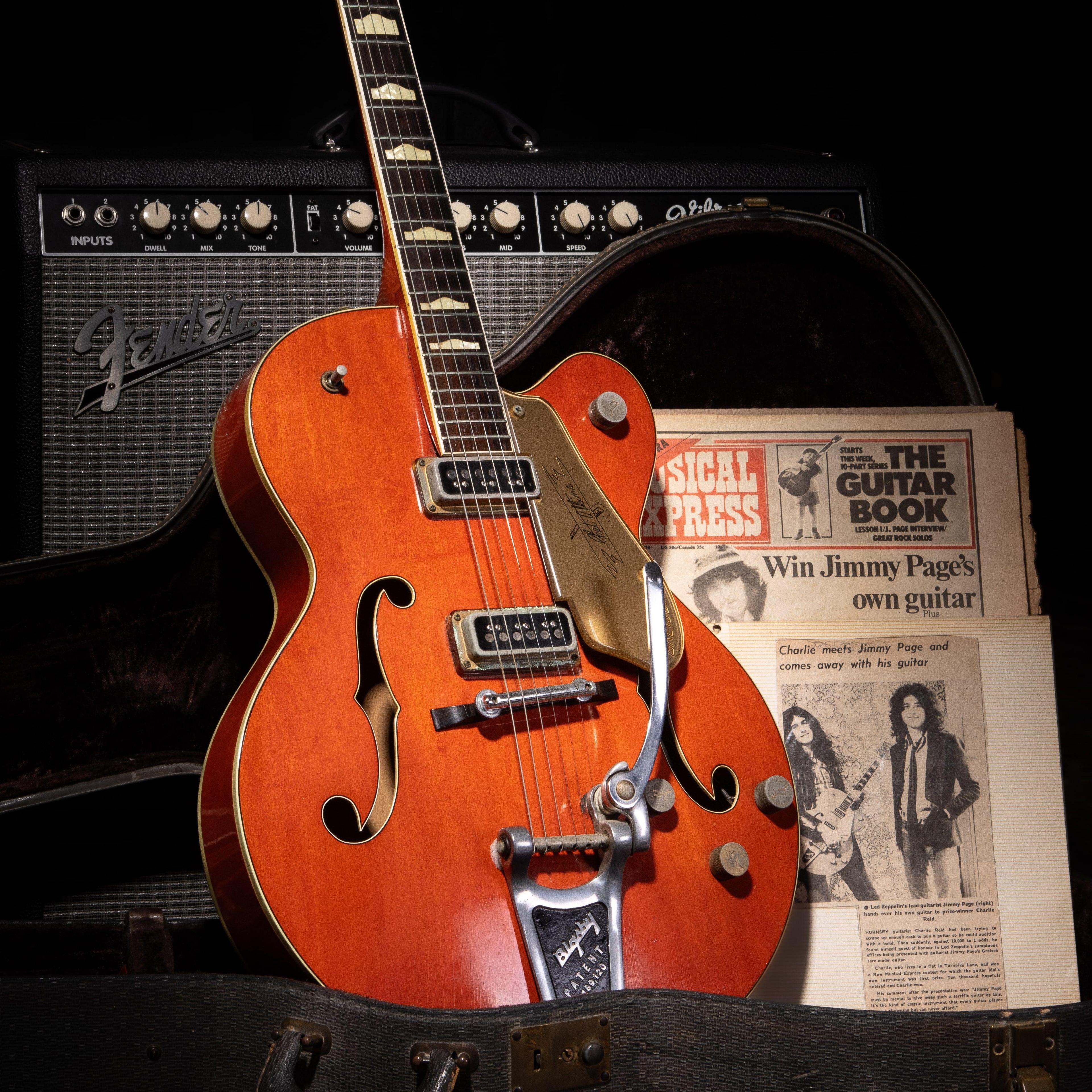 A wooden guitar leaning against an amp in a dark room. Next to it is an NME magazine in black and white, from 1974, bearing an image of Jimmy Page.