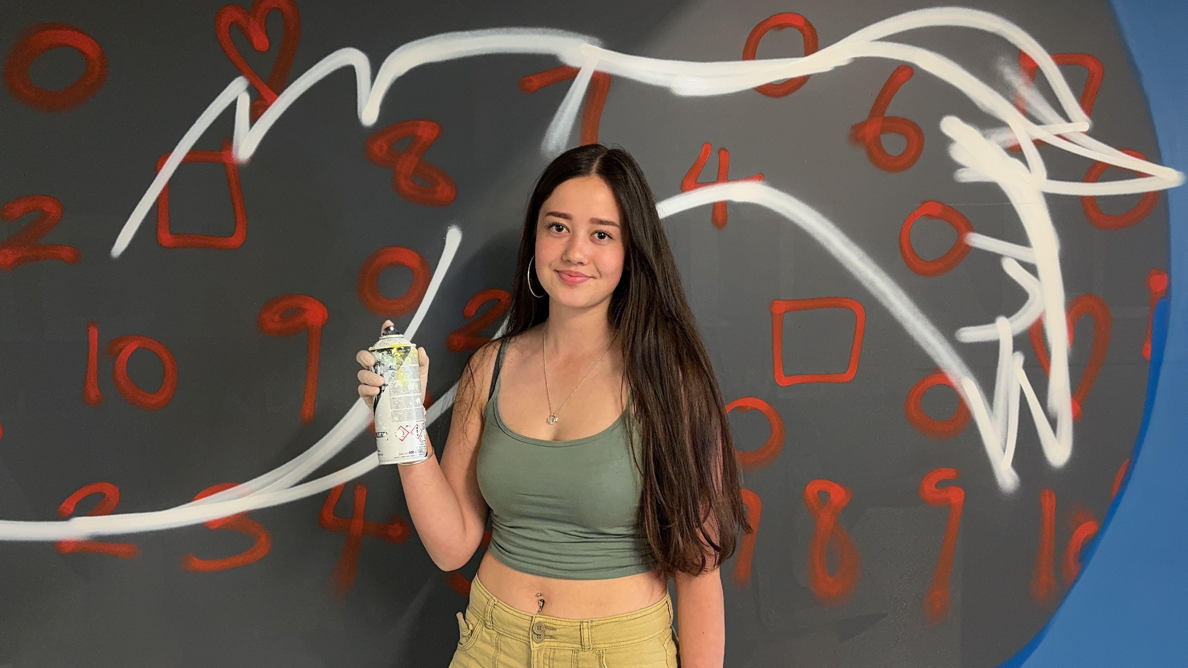 Rebecca Baldwin a young girl with brown hair stood in front of a creatively painted wall and holding a white spray paint cannister