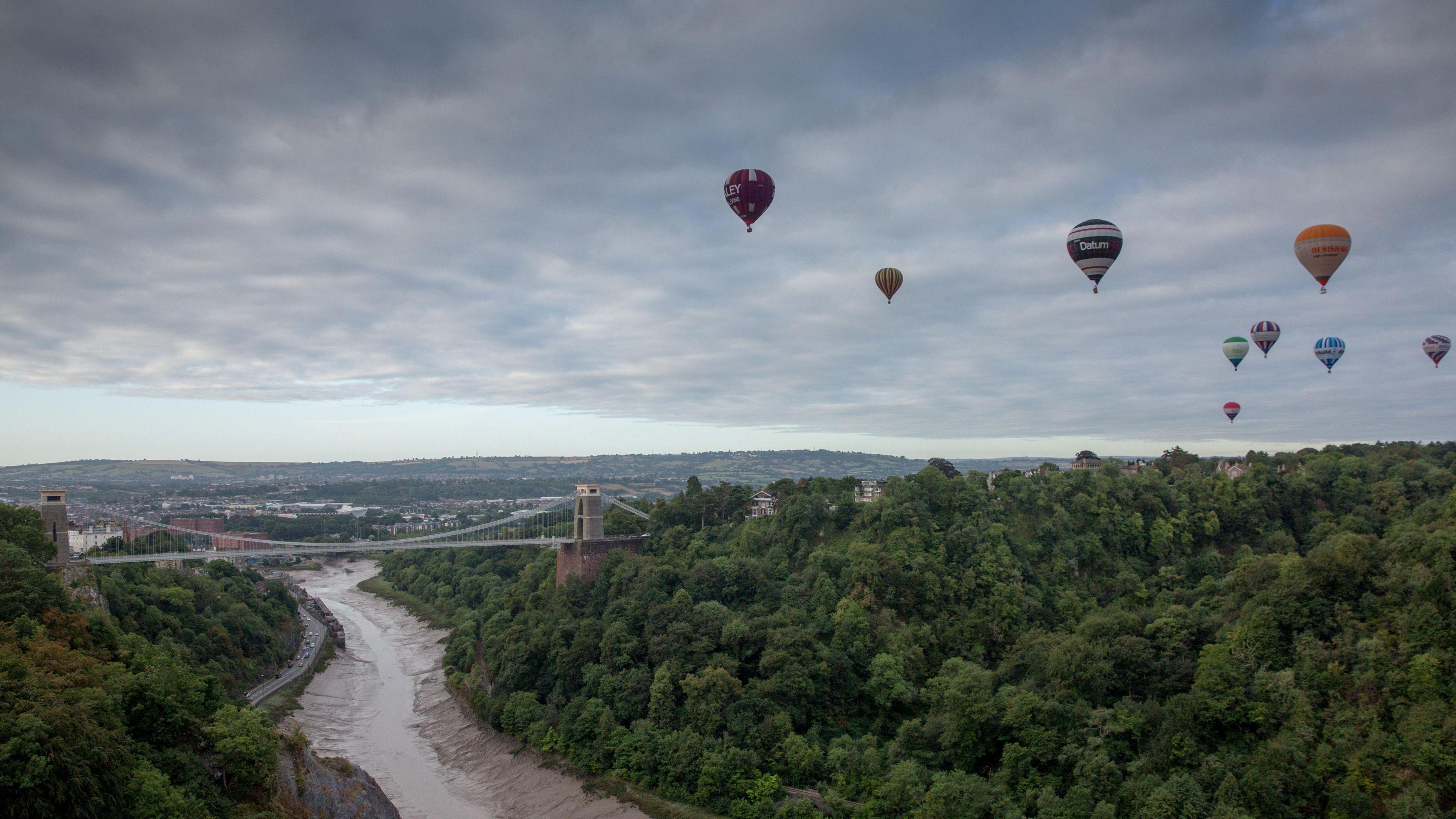 Scenic view of Clifton Suspension Bridge crossing the River Avon gorge surrounded by green forests. Several hot air balloons float in the sky above. In the background, a cityscape and distant hills are visible under a cloudy sky