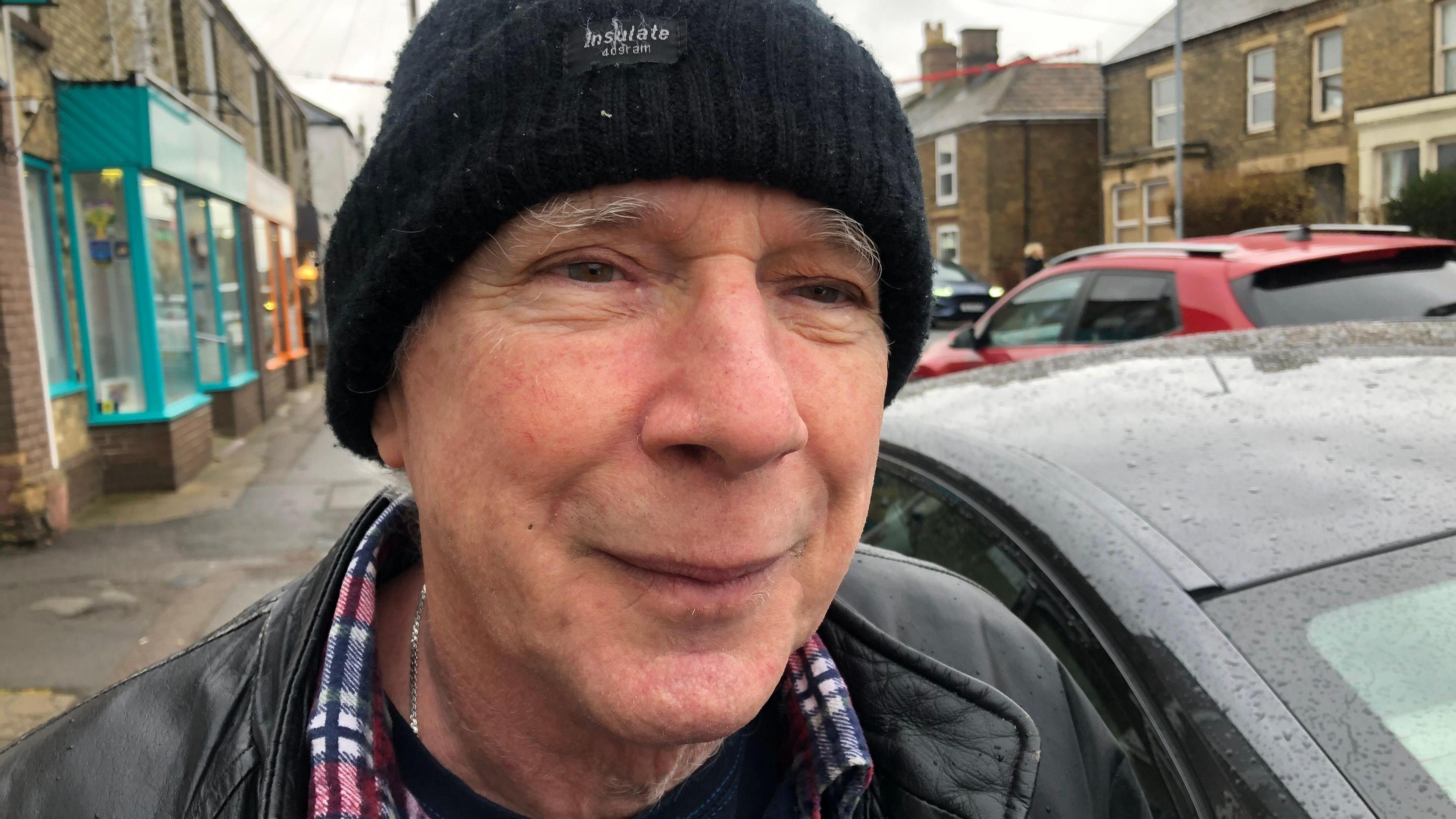 A close up of Peter Vincent wearing a black jacket, checked shirt and black beany hat. He is photographed outside with a number of cars behind him. 