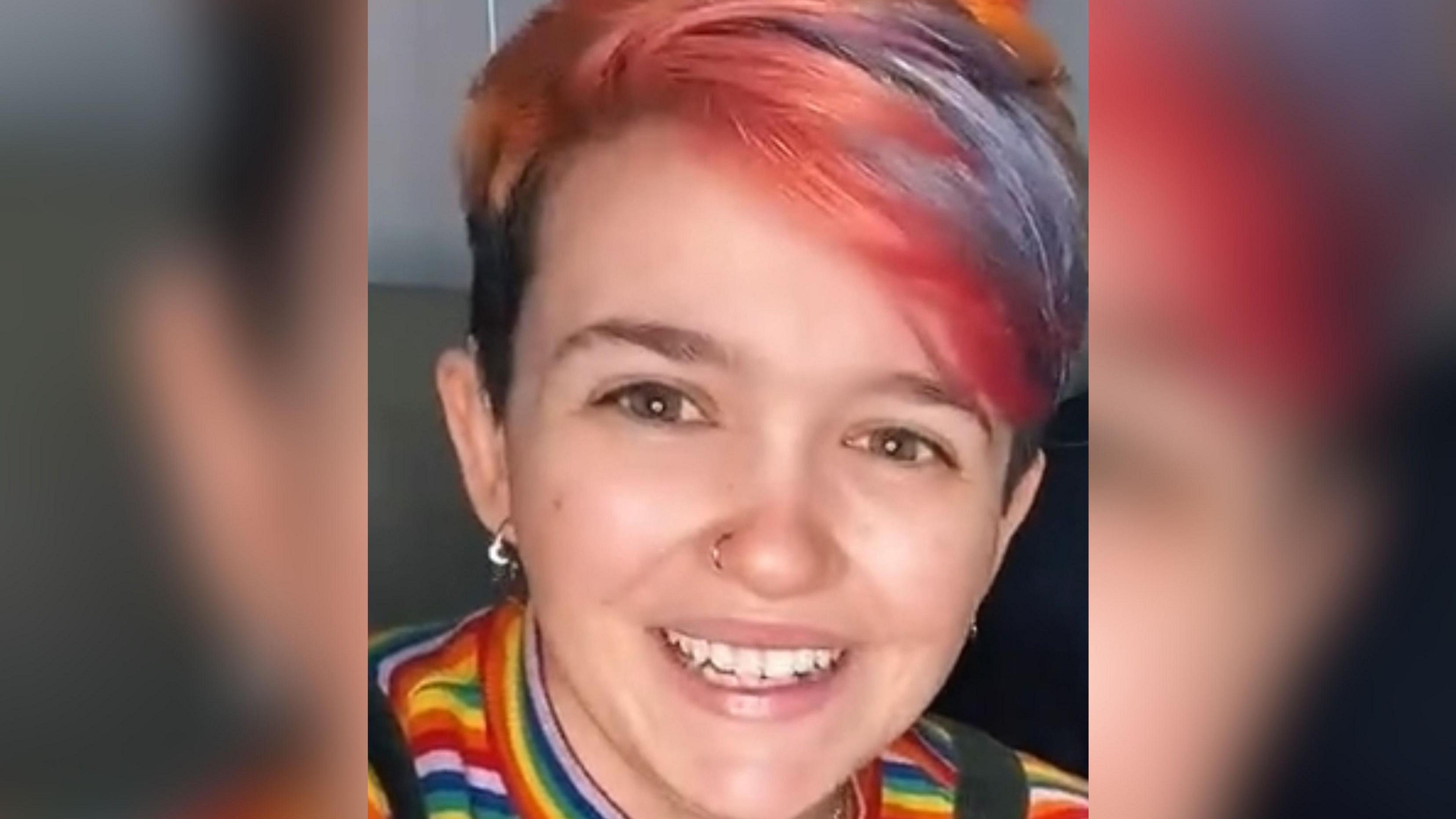 Imogen Nunn looks at the camera and smiles. She has red streaks in her hair and is wearing a rainbow striped top. Her nose is pierced.