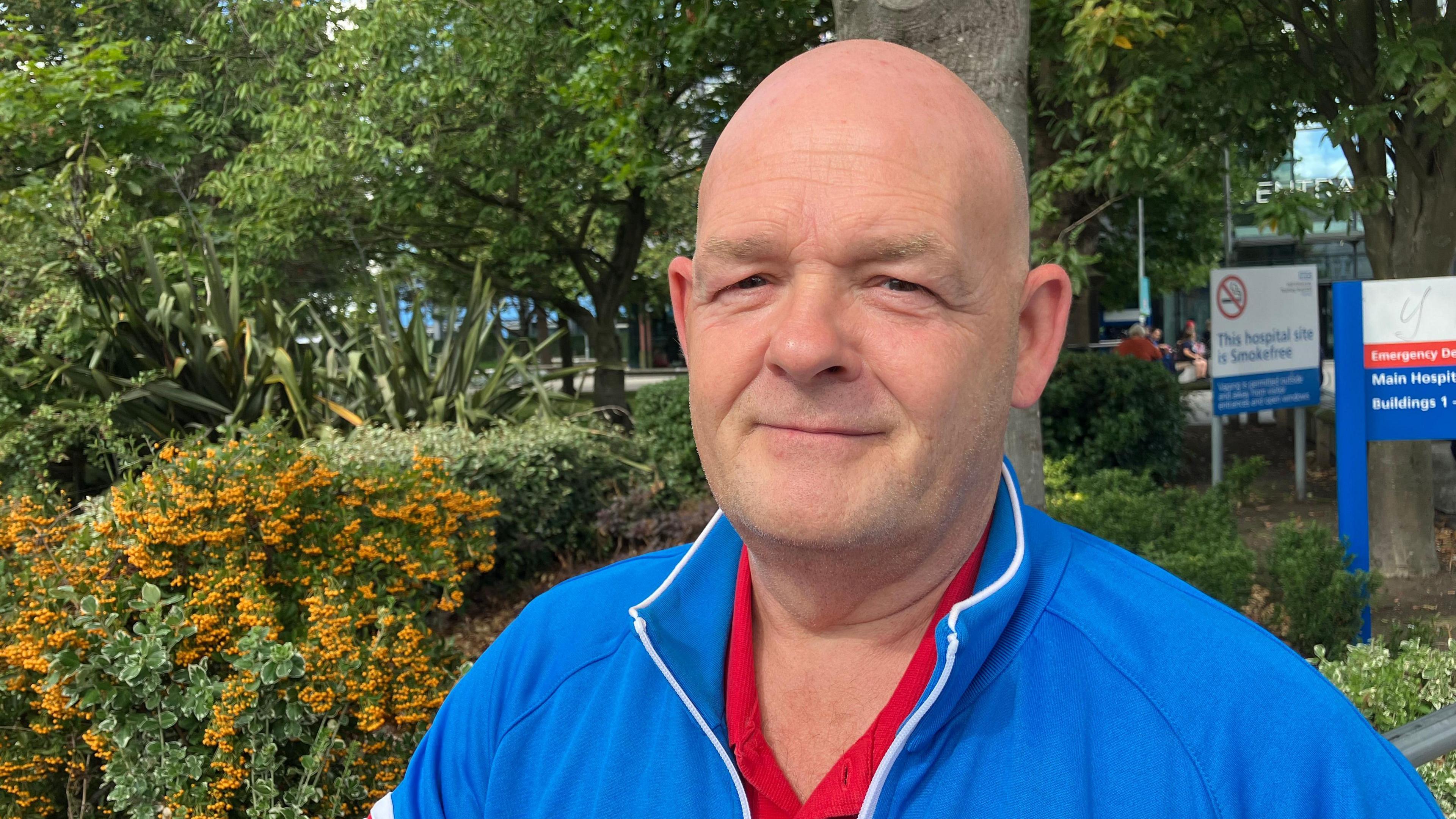 A man with a bald head is wearing a red top and blue jacket and looking at the camera smiling. You can see bushes and trees behind him and a 'main hospital' blue, white and red sign.