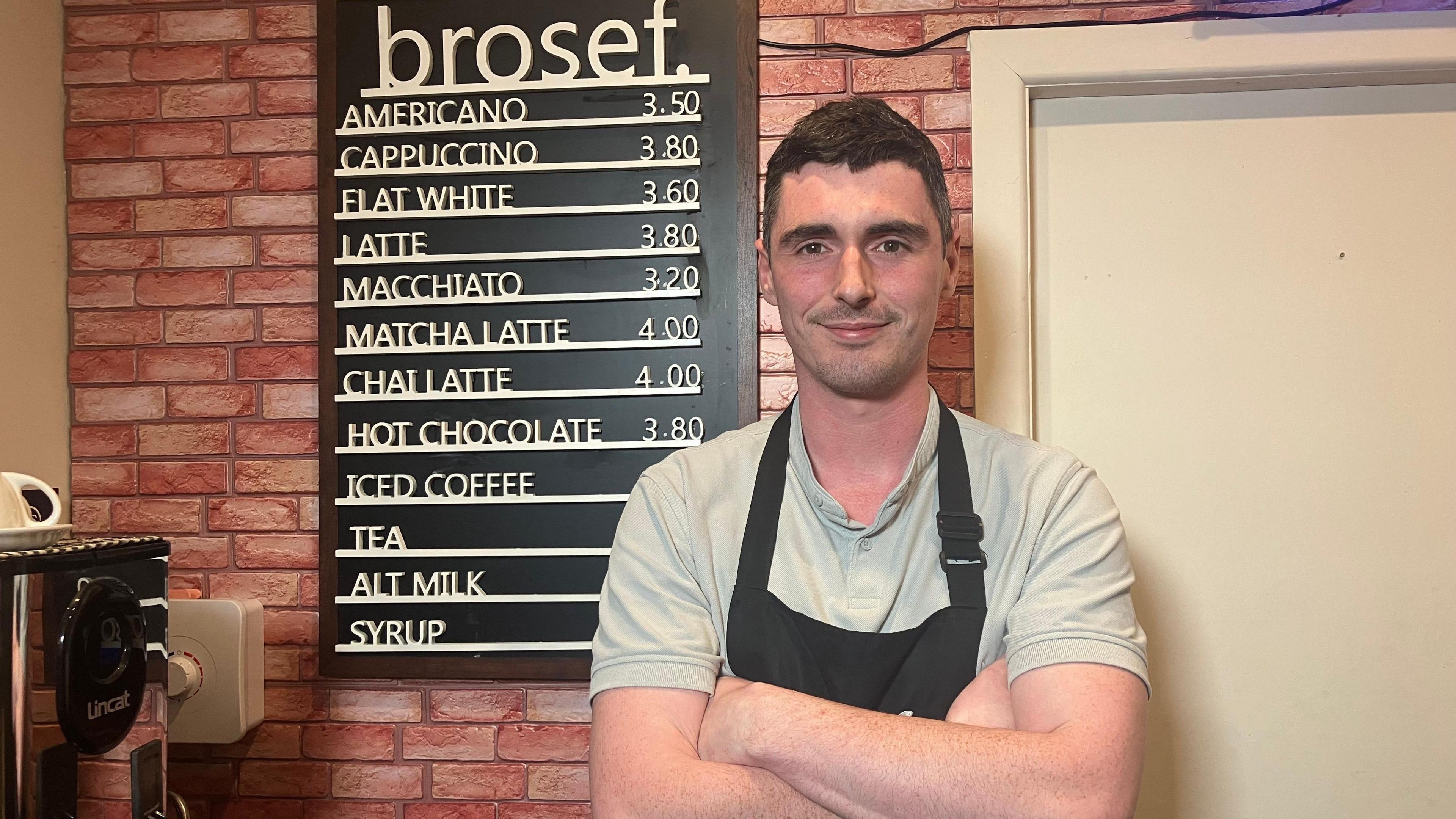 A man is standing next to a coffee menu that's attached to a wall in his café. His arms are folded and he's wearing an apron. There's a coffee machine in the corner. 