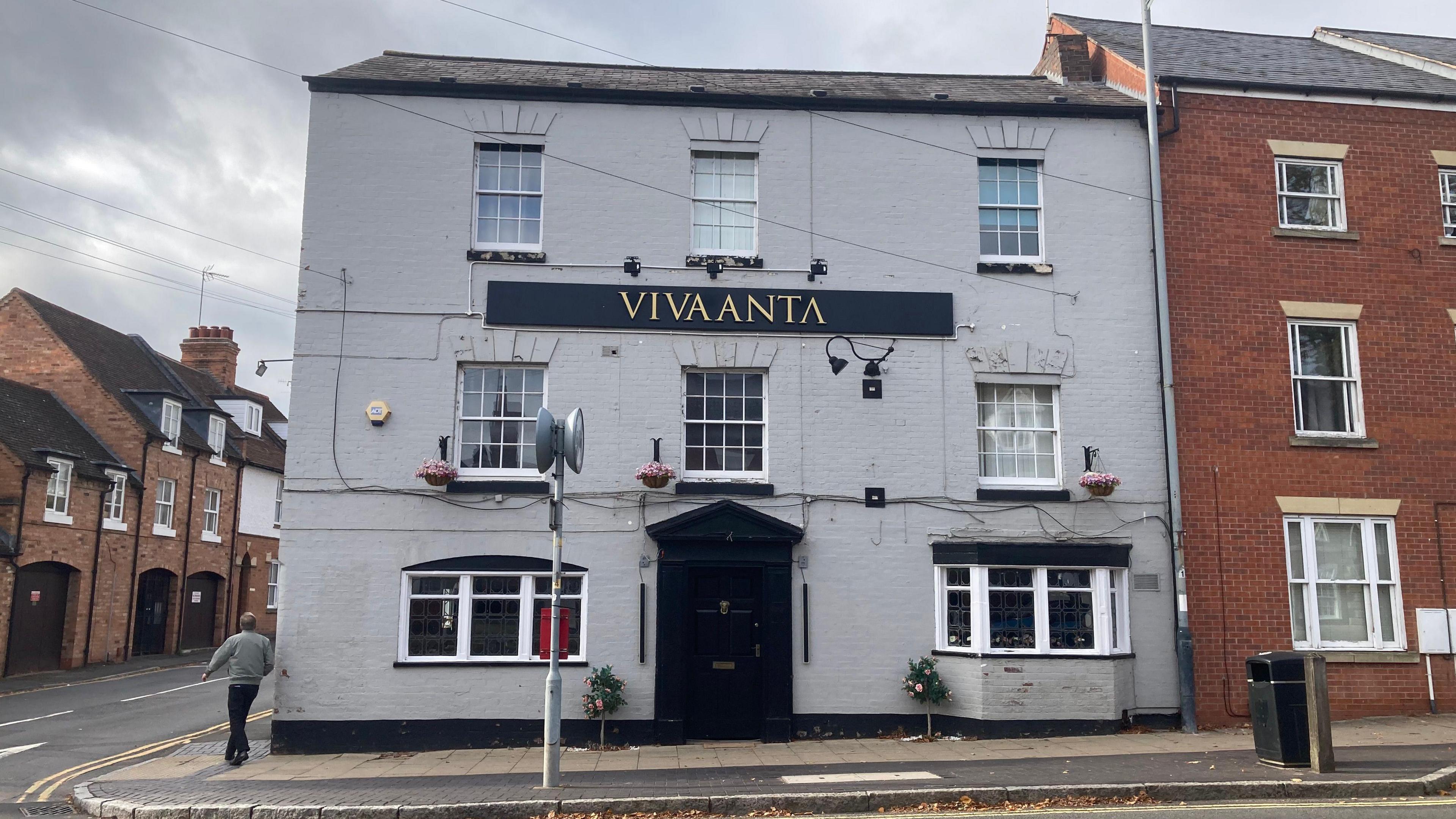 The outside of Vivaanta Indian restaurant during the day. It is a two-storey grey building on the corner of a street. It has a large black door and multiple white windows. There is a large black and gold sign on the building that reads Vivaanta. There are other properties surrounding the building.