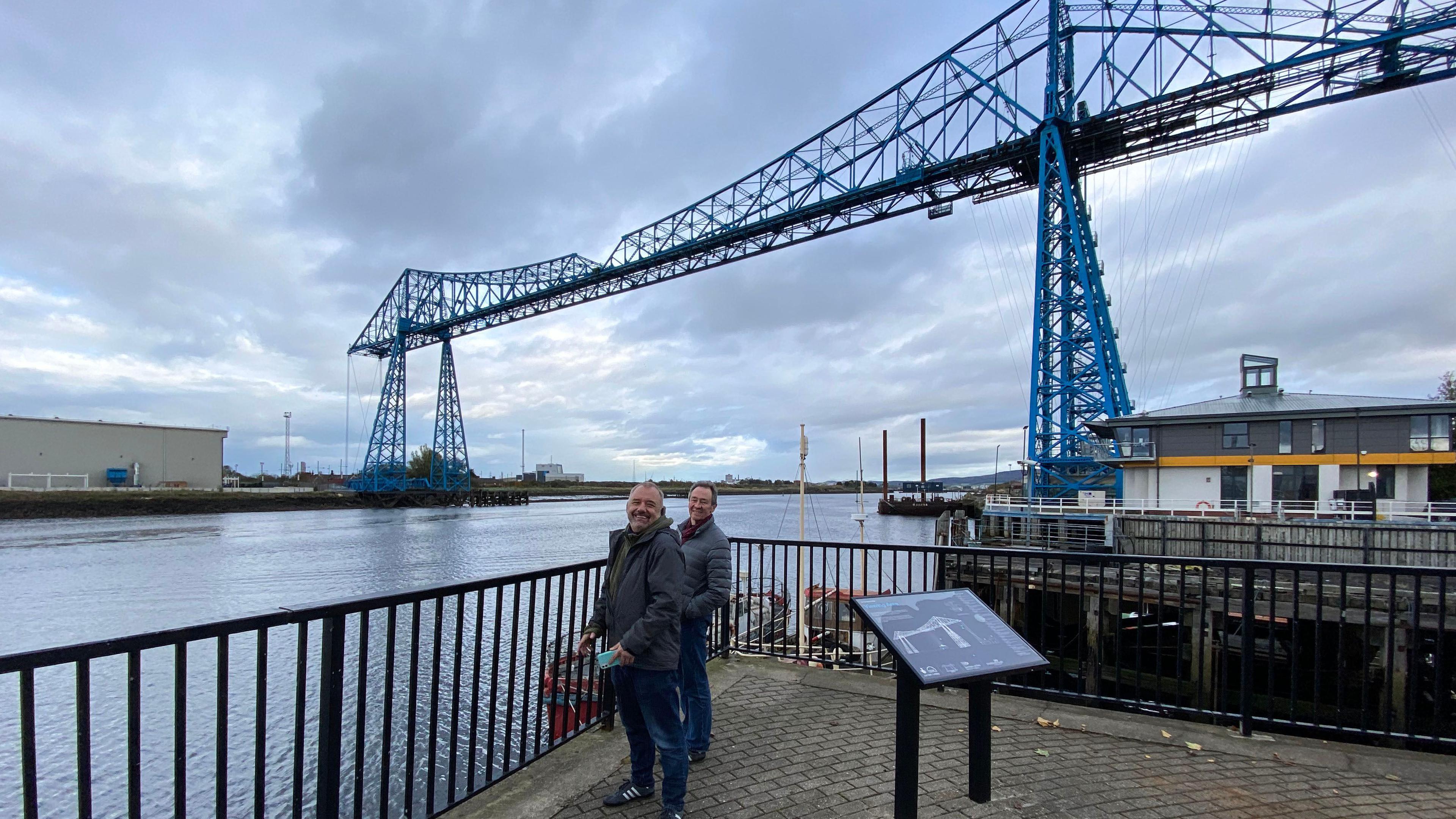 Bob Mortimer smiles at the camera, with Paul Whitehouse behind him. They are standing near a river with a metal railing in front of them. The massive blue Transporter Bridge is behind them. It is a cloudy day. 