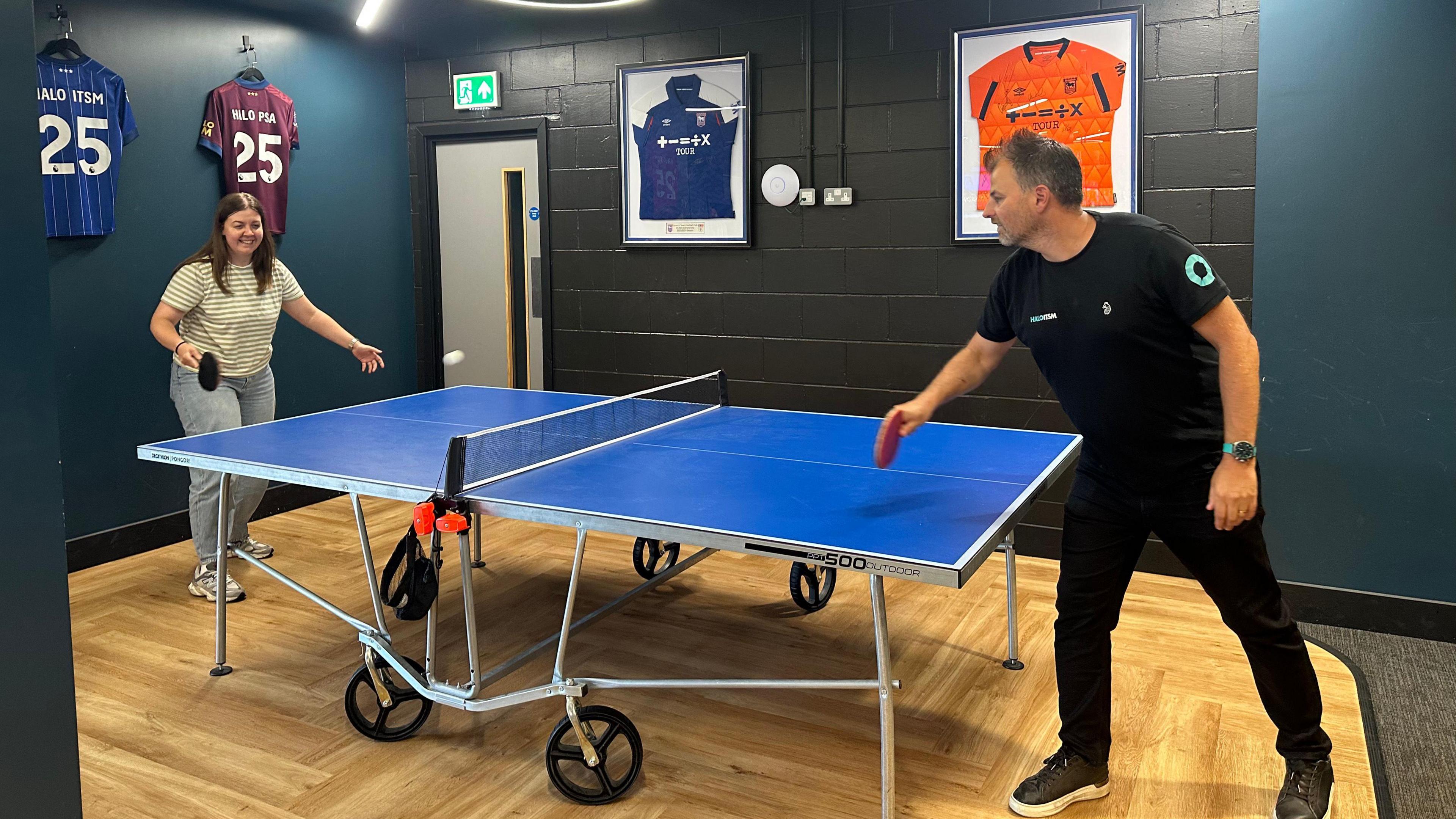 Mr Hamilton plays table tennis with one of his staff members in the Halo officers. Framed Ipswich Town shirts have been hung on the wall behind the ping pong table. 