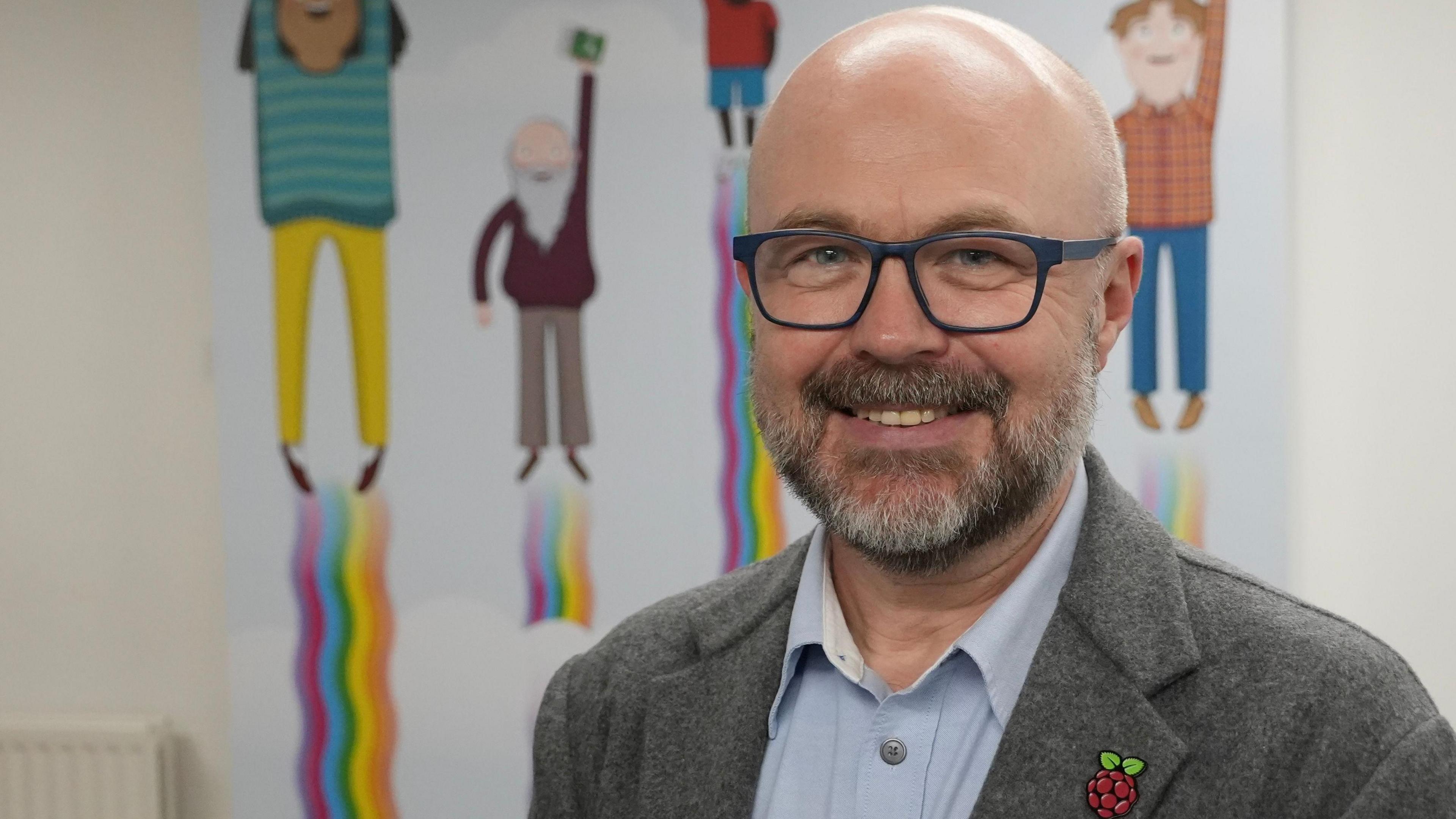 Philip Colligan has glasses and a beard and is dressed in a grey jacket and light blue shirt. He is standing indoors in front of a colourful illustration. His jacket features a Raspberry Pi logo pin. Behind him, a vibrant poster shows four cartoon characters standing on rainbow-colored beams, each holding a small green item.