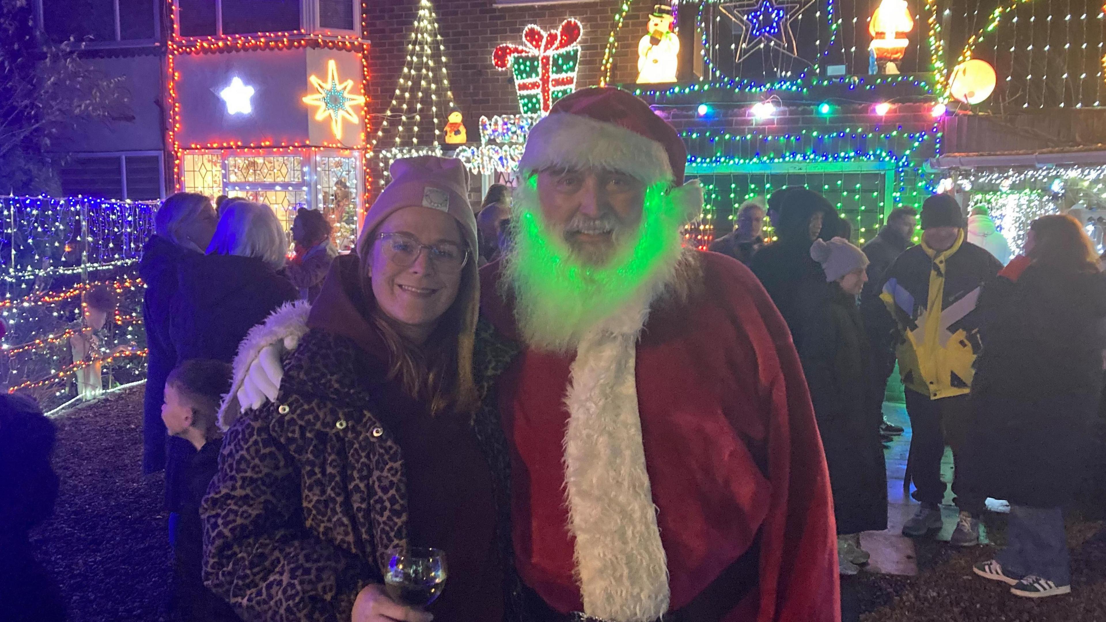 Siobhan Kirby, wearing a pink hat and a leopard print coat, stands next to her uncle Anthony O'Toole, who is dressed as Father Christmas with a ring of green lights in his beard. Behind them, Mr O'Toole's house is bedecked with lights and people have gathered to admire them.