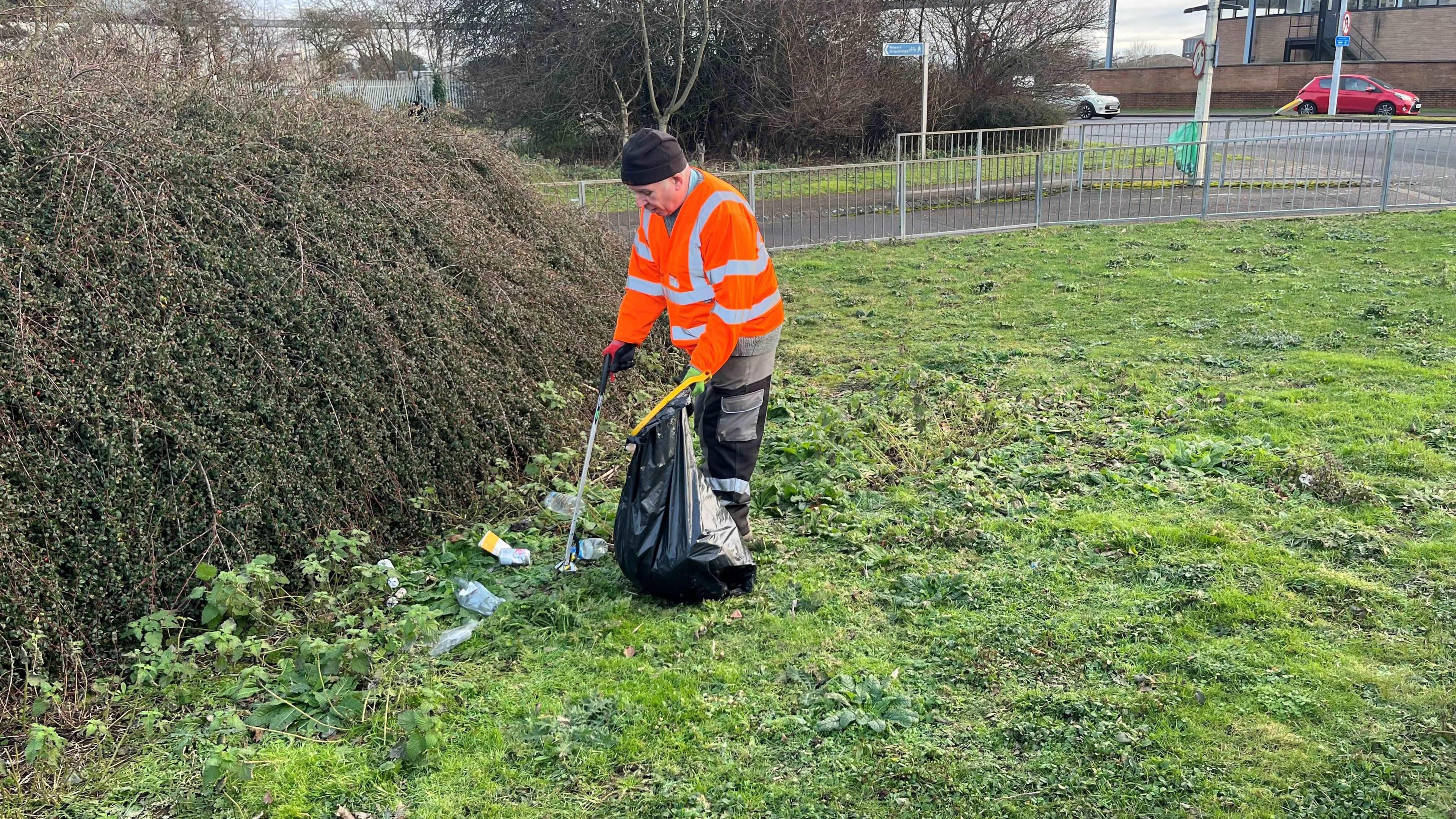 Mark Fishpool, 68, has a black beany hat on with some grey hair and side burns visible. He is wearing a Hi-Viz orange jacket with a grey jumper underneath, he has red gloves and is holding a litter grabber in one hand and a black bin liner in the other, there is a hedge in front of him, he is collecting plastic bottles, paper cups and crisp packets from a grassy are near a dual-carriageway.