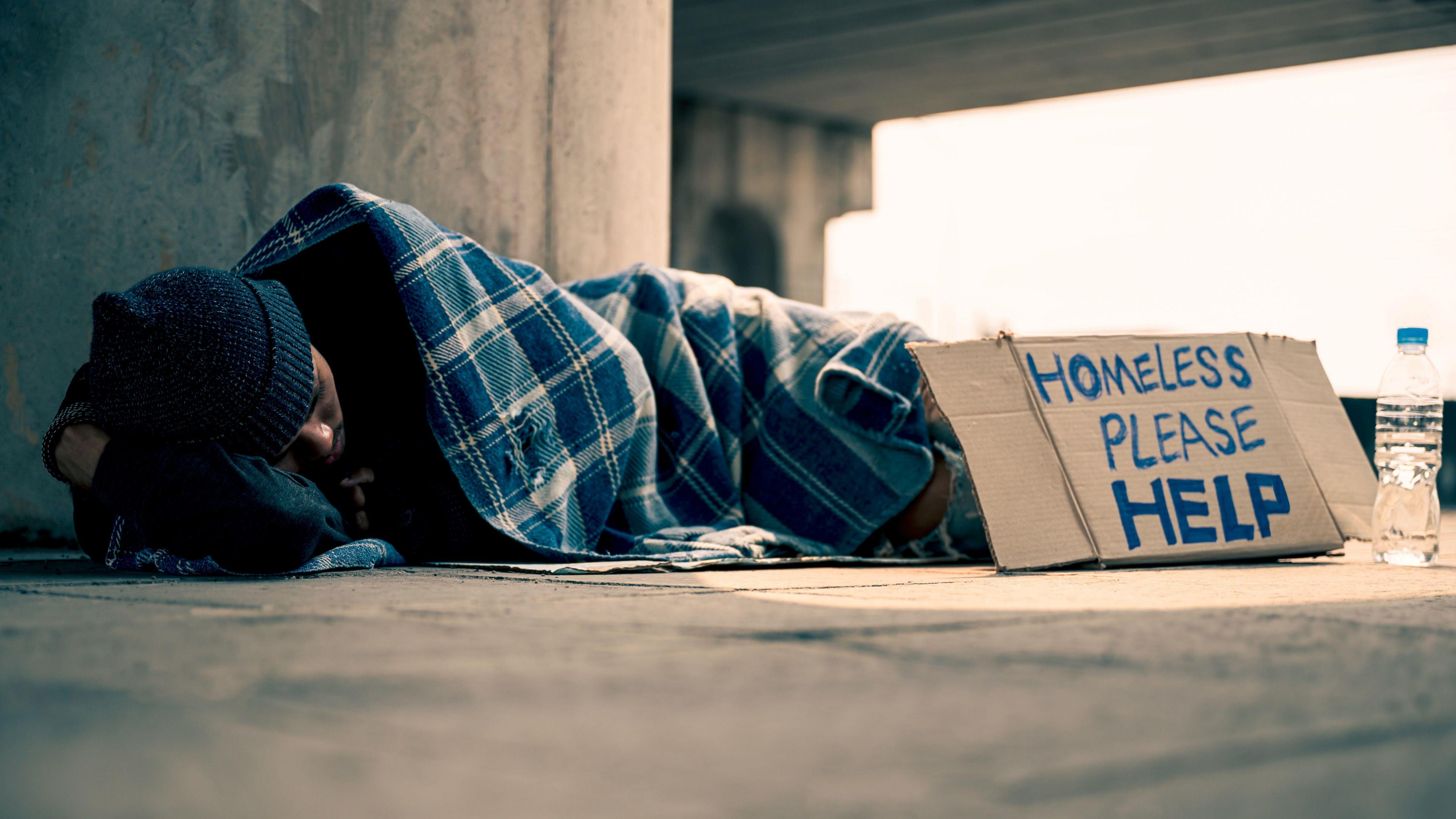 A man lies sleeping under a blanket with a woolly hat in a street. He has a handwritten cardboard signs that reads "Homeless, please help".