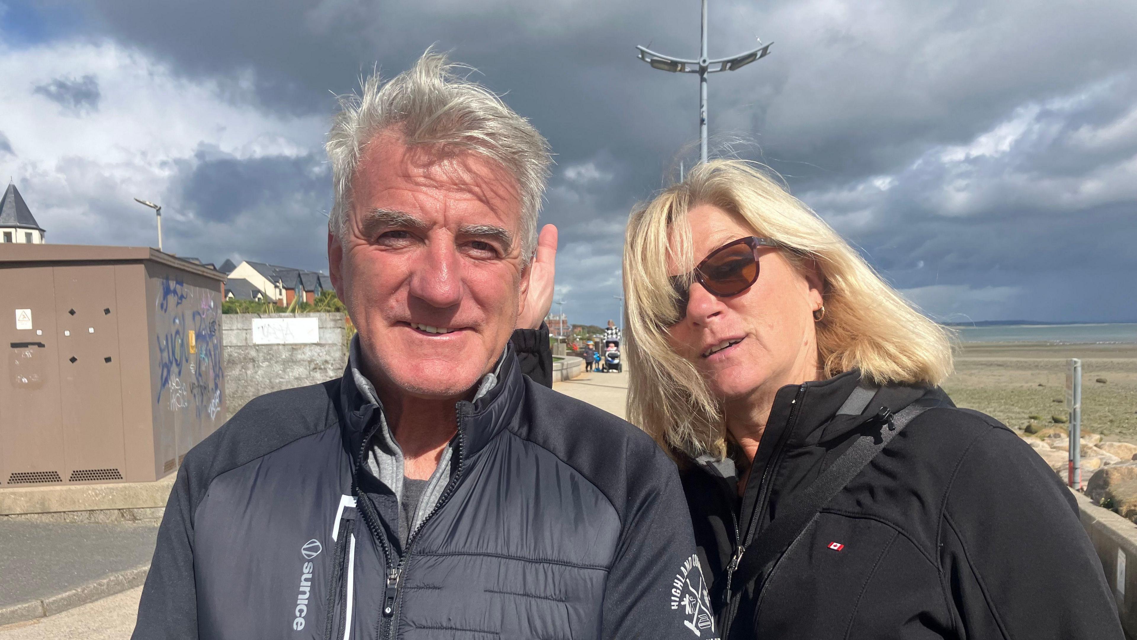 Ted is wearing a grey puff jacket. He is smiling at the camera and has grey hair and a tan.

Julie is mid pose with blonde hair and brown sun glasses. She is wearing a black ski jacket. 

You can see the beach behind and the sea. The sun is shining. 