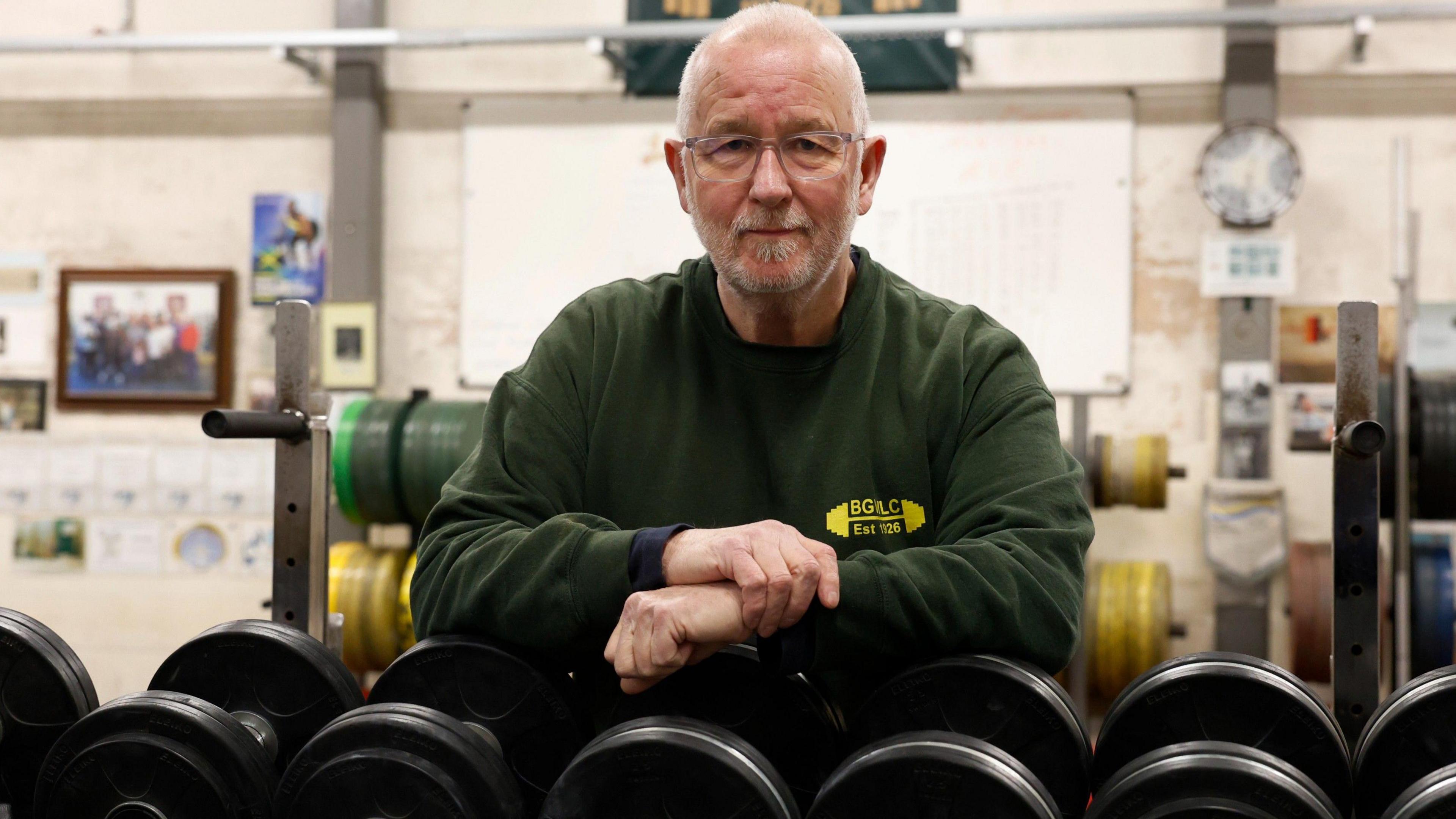 Martin Bass is seen leaning on a rack of free weights facing forward. He is wearing a green jumper with the club's logo on it.