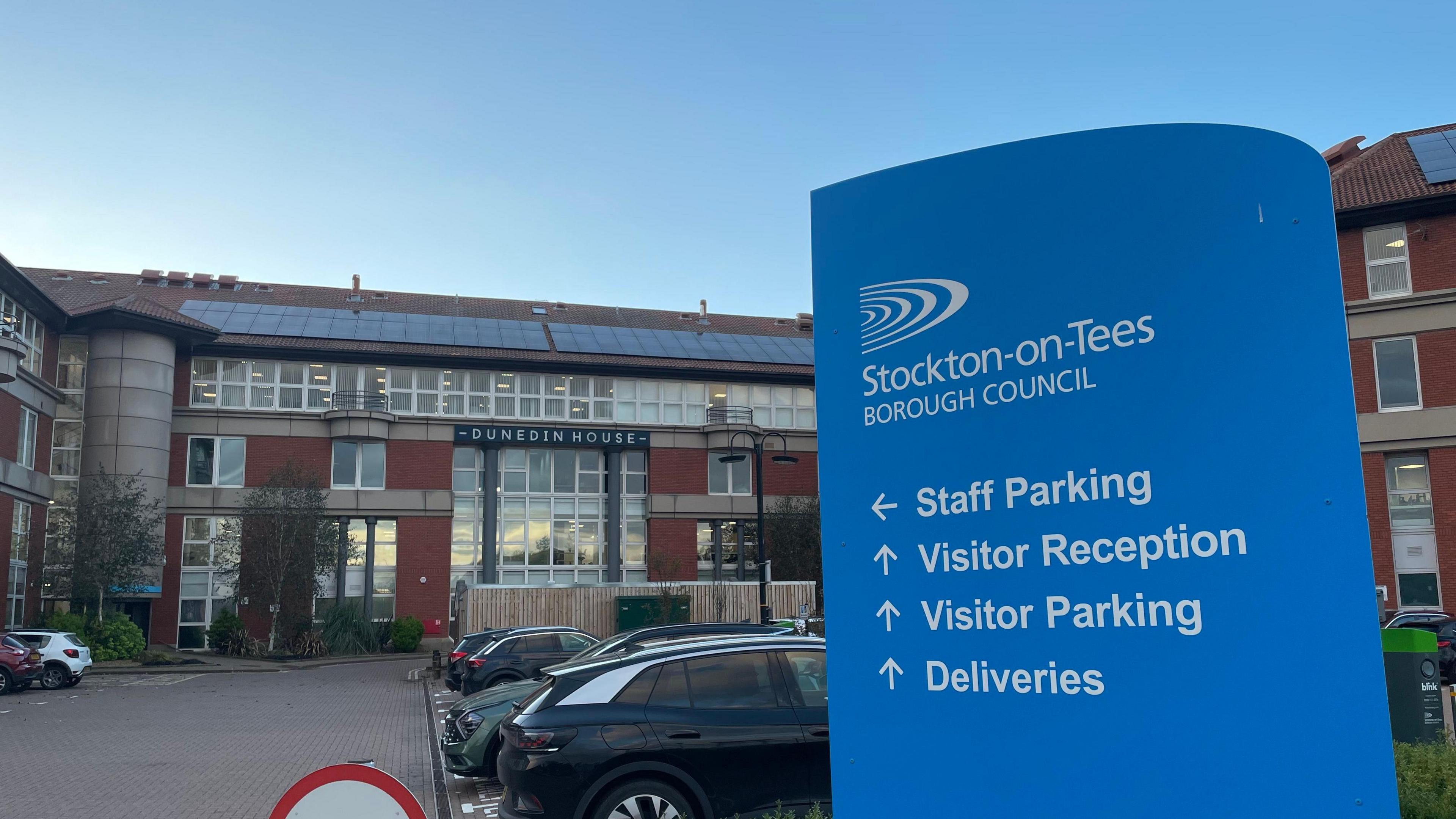 A blue sign that reads 'Stockton-on-Tees Borough Council, with staff parking, visitor reception, visitor parking and deliveries in lettering underneath. Behind the sign is a large red brick building with a car park in front of it. 