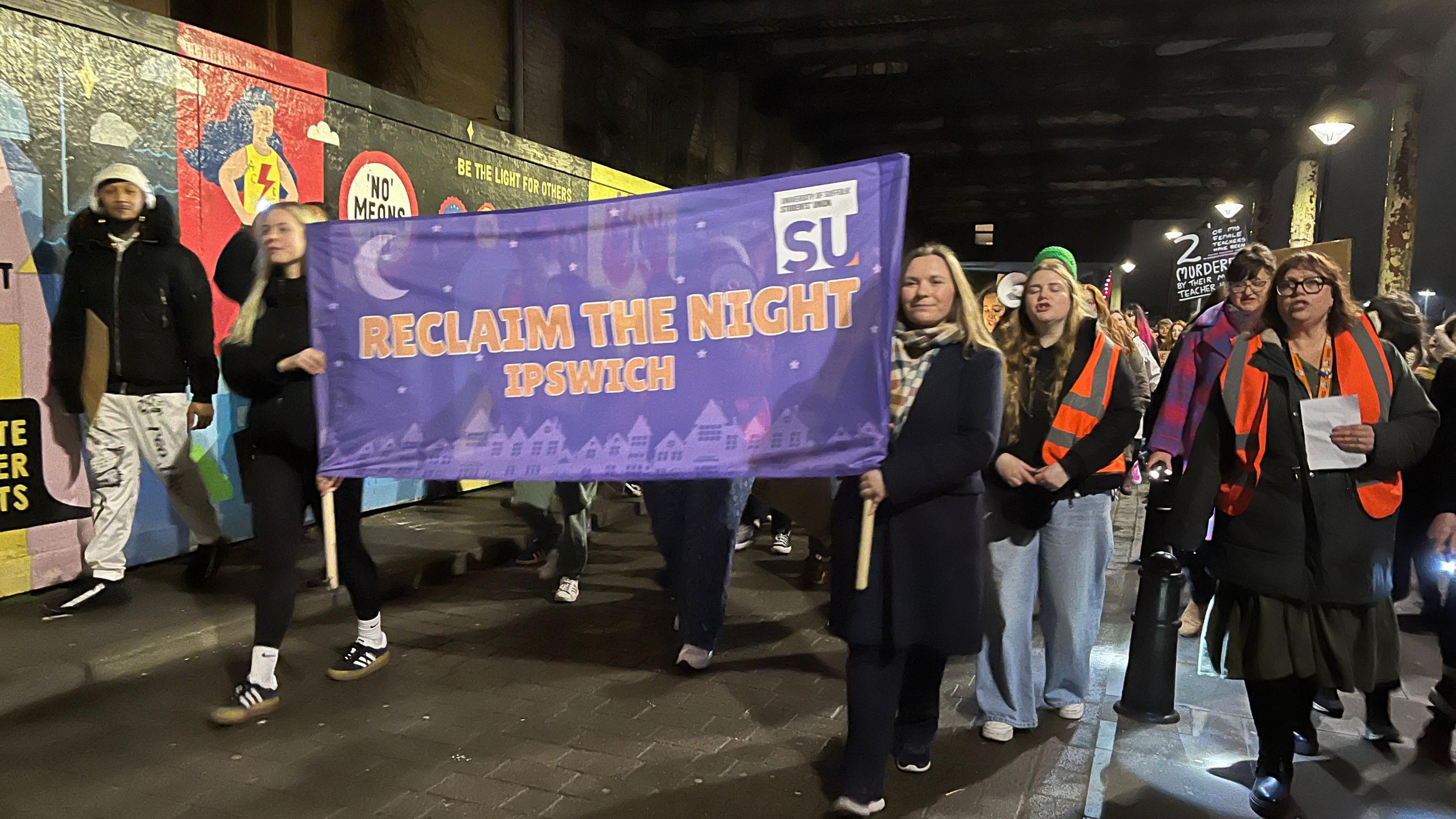 Two women holding a banner which reads Reclaim the Night walk along a street. They are flanked by crowds of men and women walking beside them as part of a demonstration.