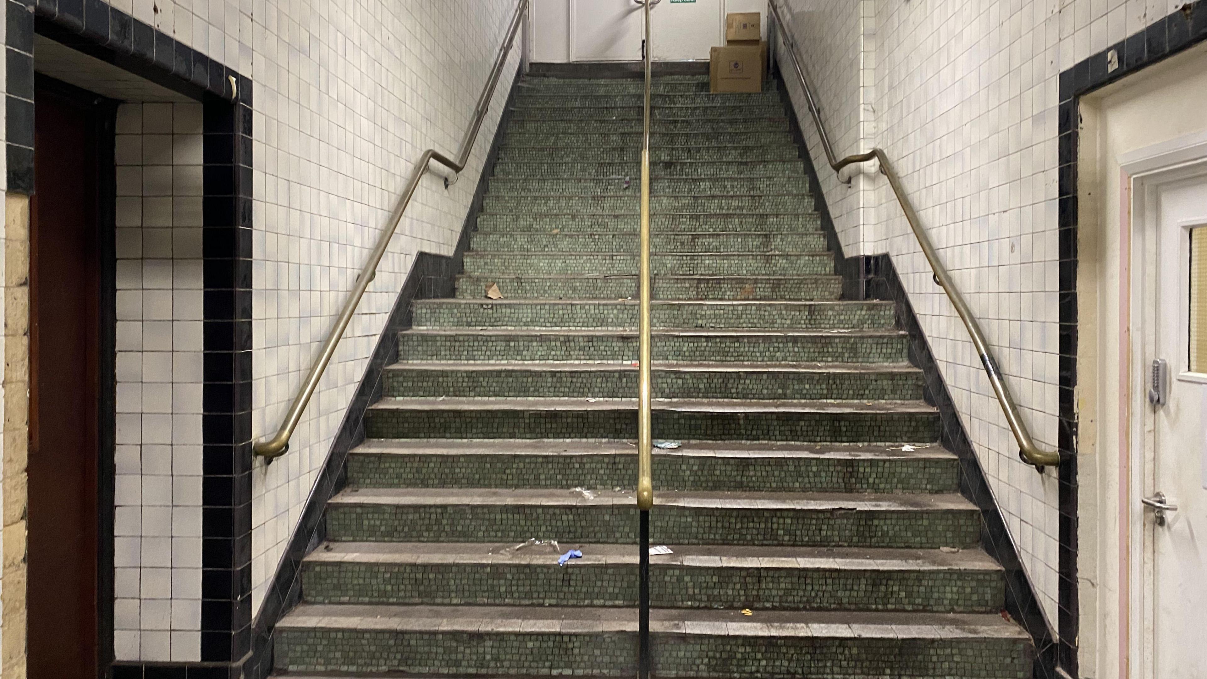 A stairwell, the stairs a turquoise but they are covered in dirt. There are white and black tiles on the walls. The banisters are golden. 