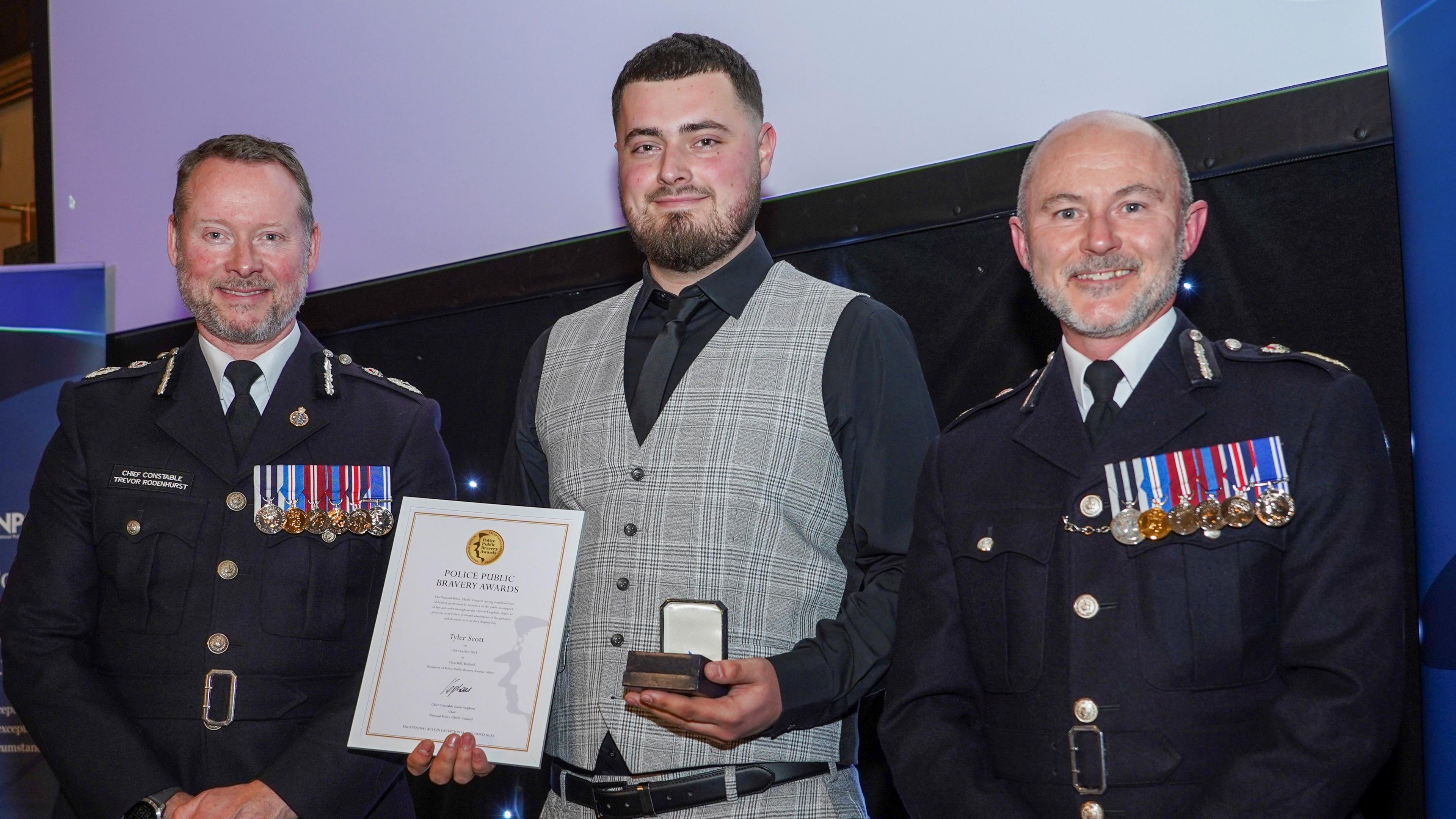 A man with dark brown hair and short beard, wearing a grey waistcoat and black shirt and tie, holding a police bravery award certificate and a medal in the other hand. He is flanked on either side by two senior police officers.