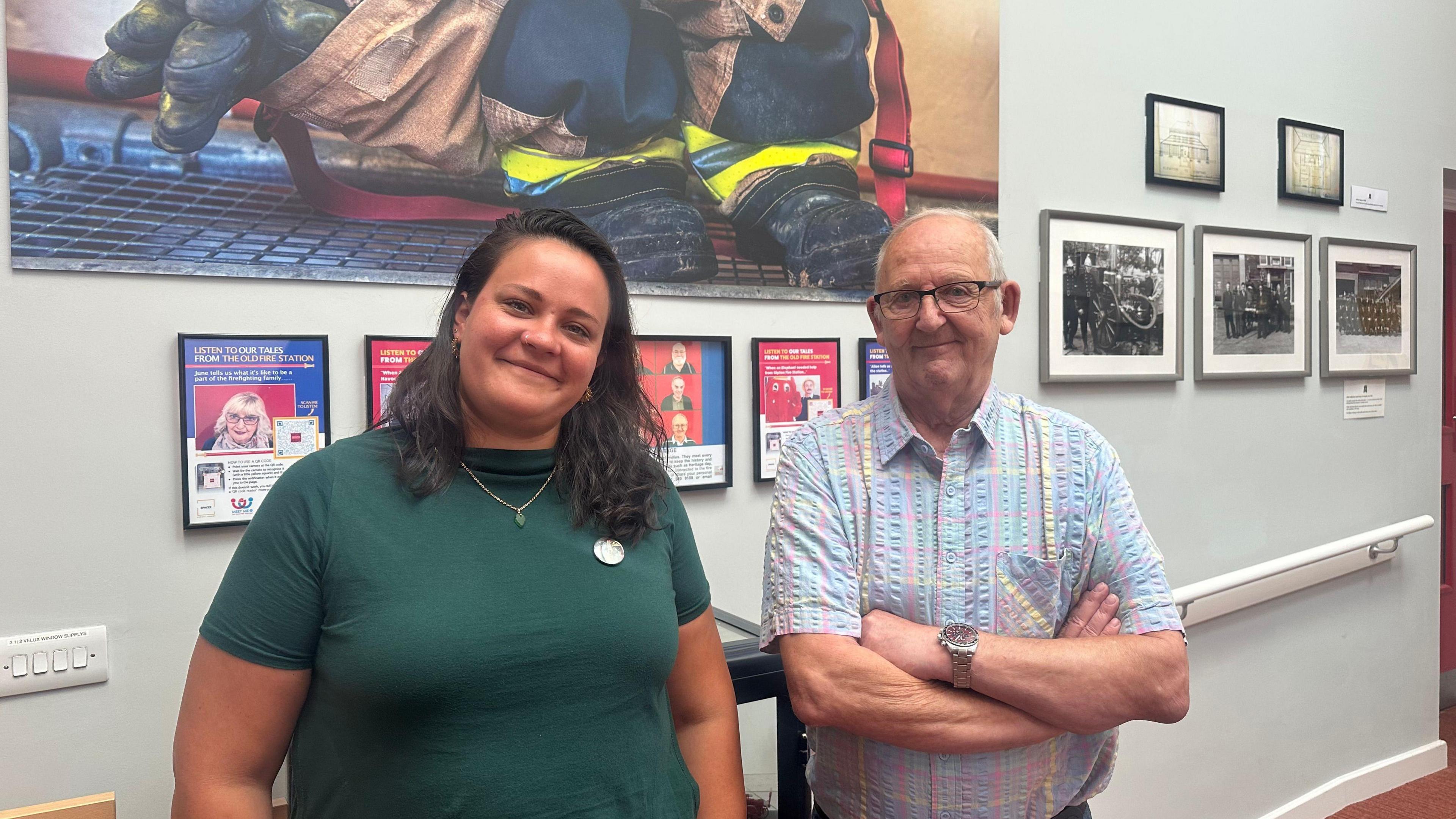 A lady with dark hair wearing a dark green t-shirt smiles stood next to a man in his 80s who is stood with his arms folded. He is wearing a light coloured checked shirt and they are both stood in front of a wall that has old fire station pictures on the wall.