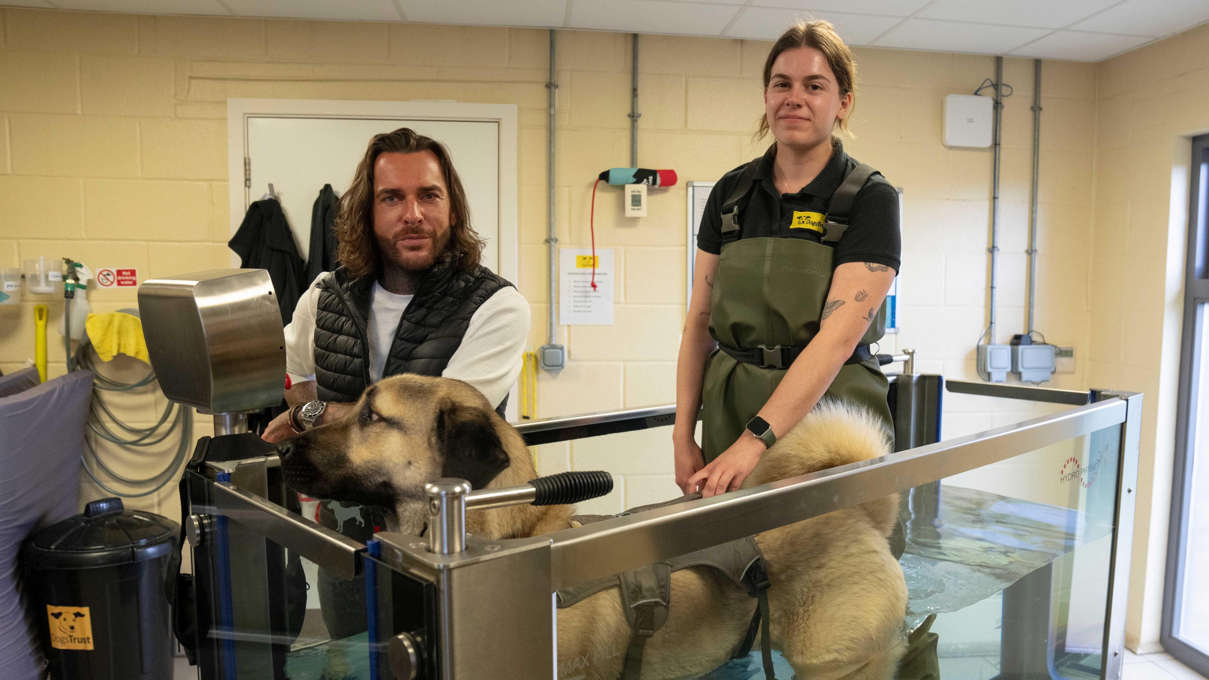 A man, Pete Wicks, stands next to a canine hydrotherapy unit in which a large dog is standing in water up to its belly. A woman wearing a thick apron stands next to the tank as well.