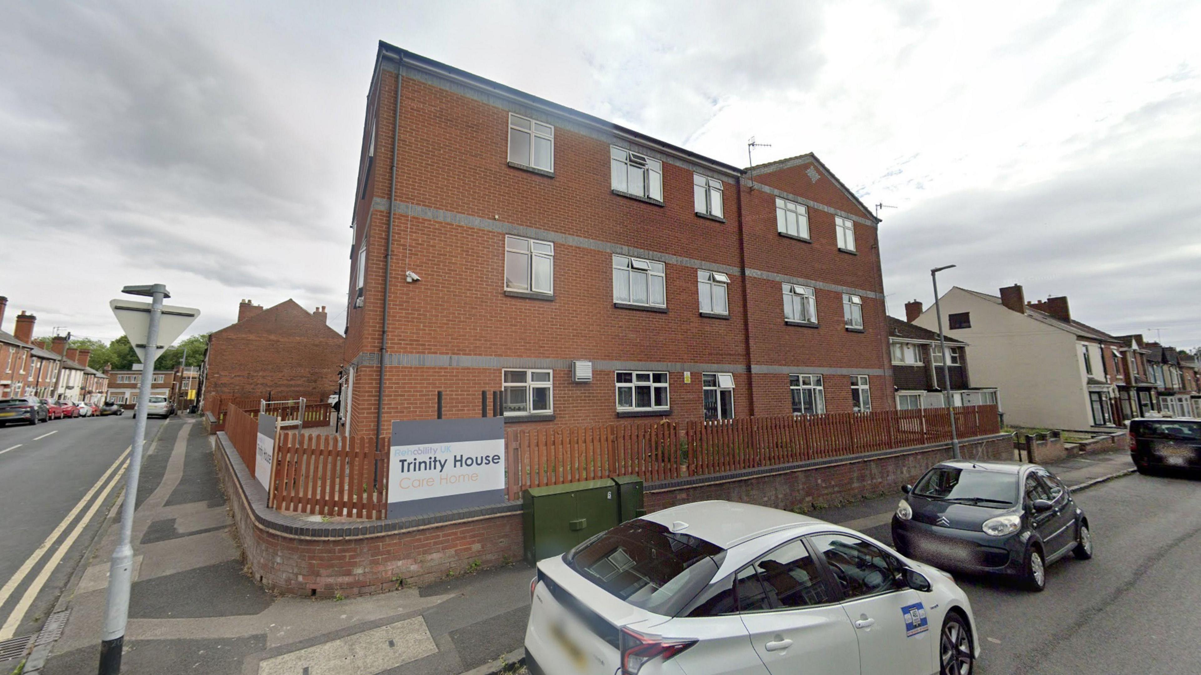 A three-storey, red-brick building on a corner of two streets. There is a low wooden fence around the building. There is a white sign on the fence which reads "Trinity House Care Home".