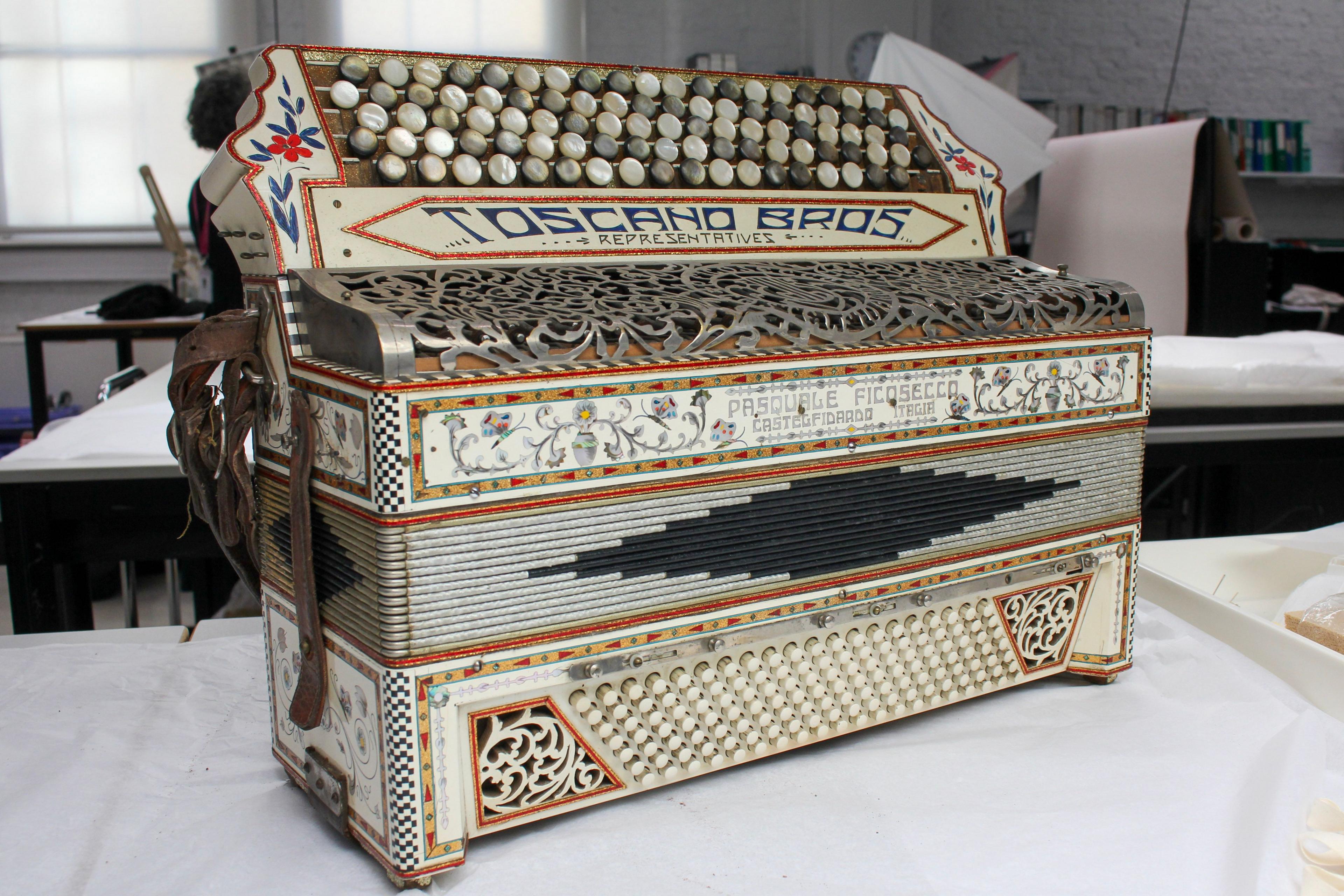 An old-fashioned white brass accordion on a white-cloth table. The accordion has red and white floral decorations on it and 'Toscano Brothers and representatives' is written along it.