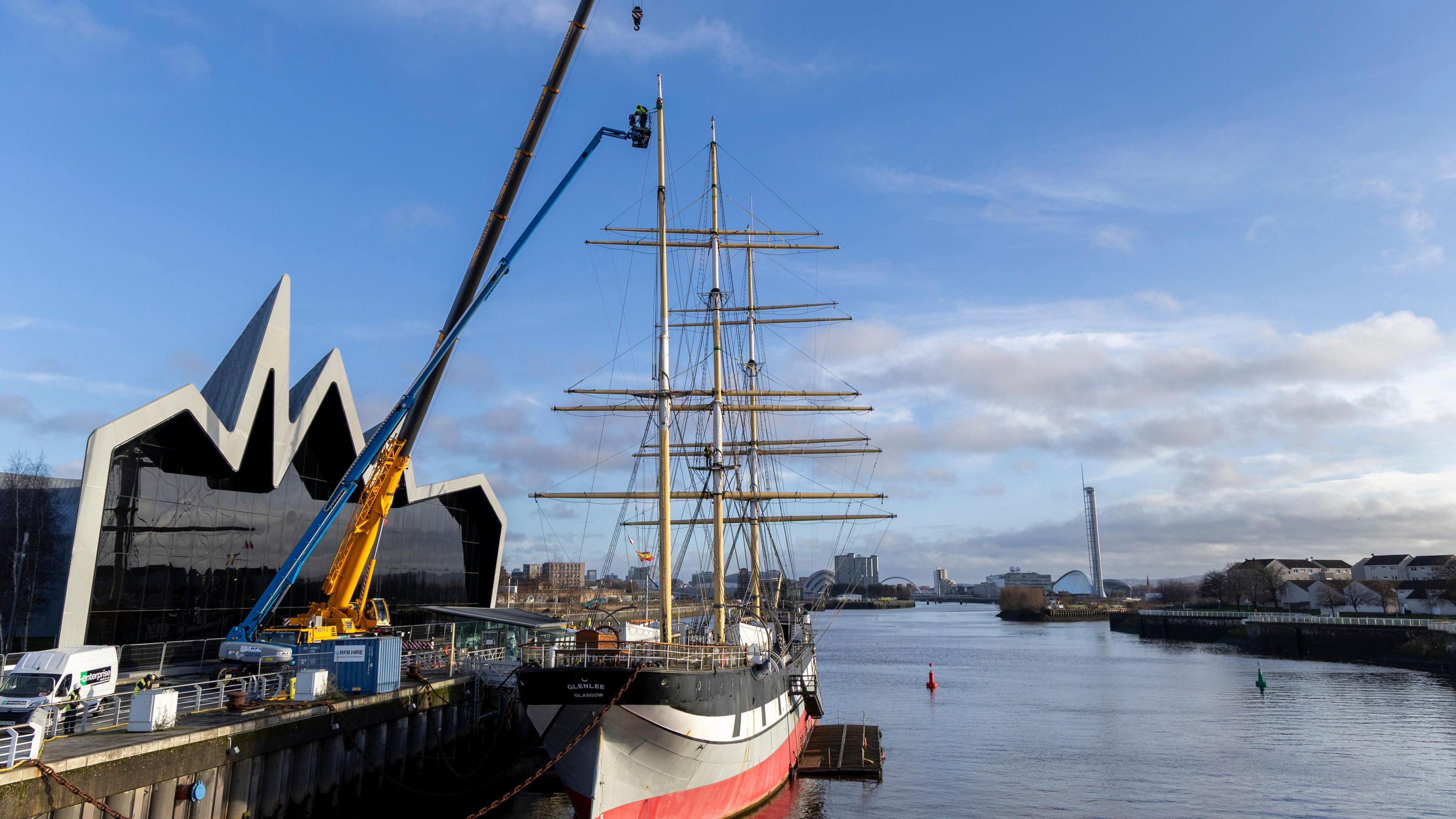 A sailing ship docked next to a large museum building. A large crane and other construction equipment are next to it, with wok clearly taking place on the boat.