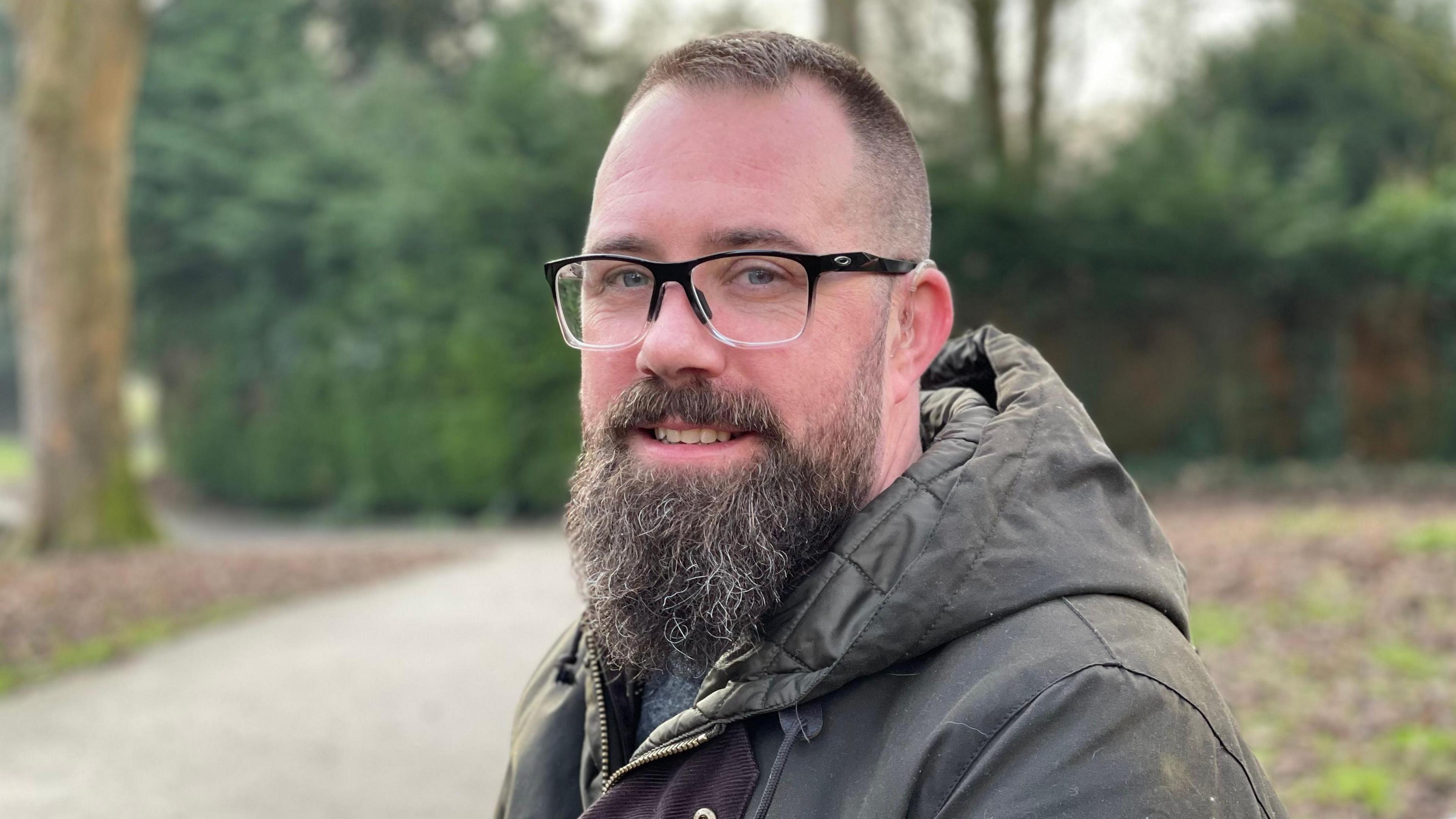 Photo of man with a long beard and green waxed coat with hood, wearing glasses, smiling, in a park with trees and bushes behind him