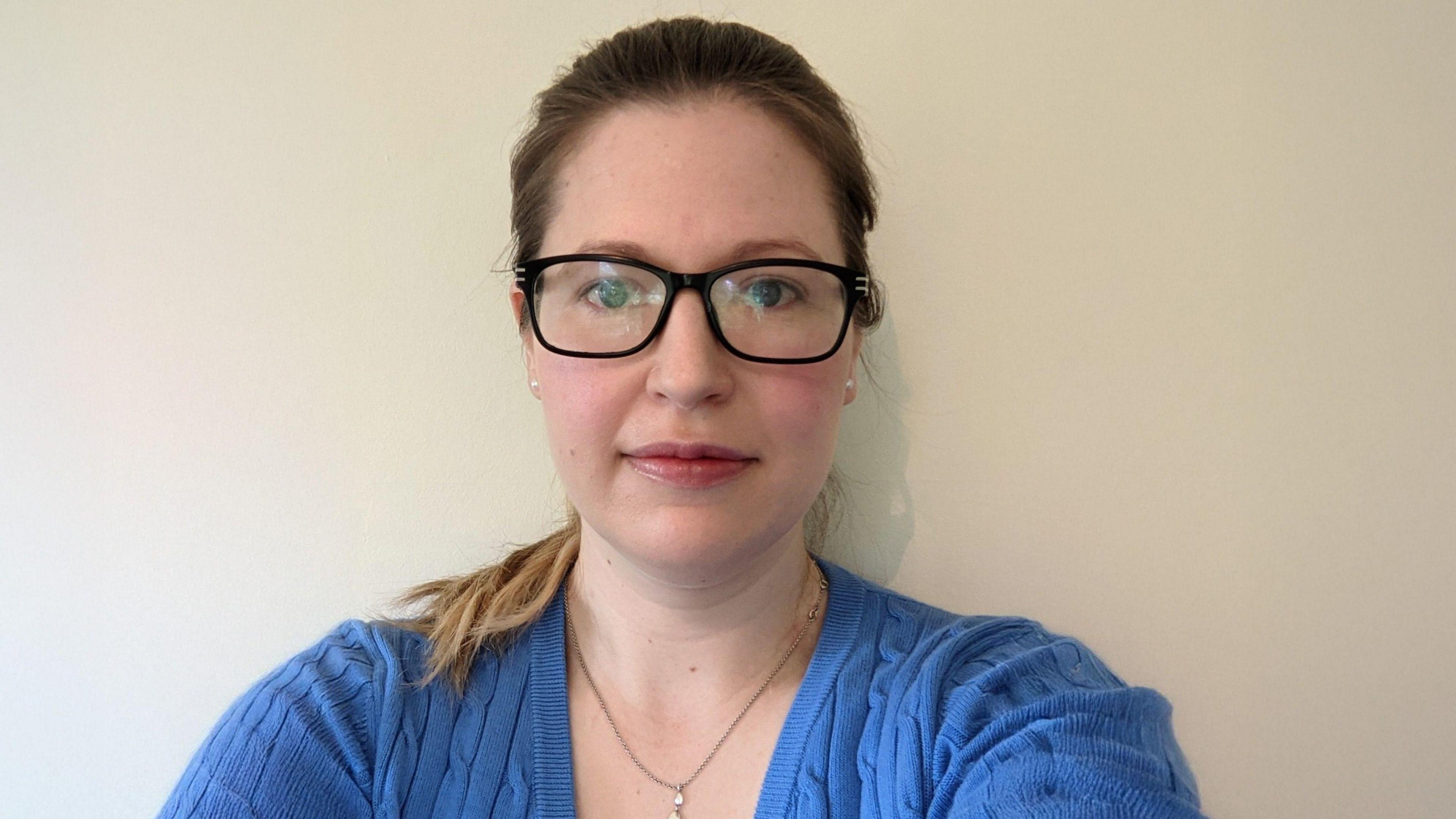 A woman with her hair tied back wearing a blue jumper and black glasses looking directly at the camera and smiling slightly.