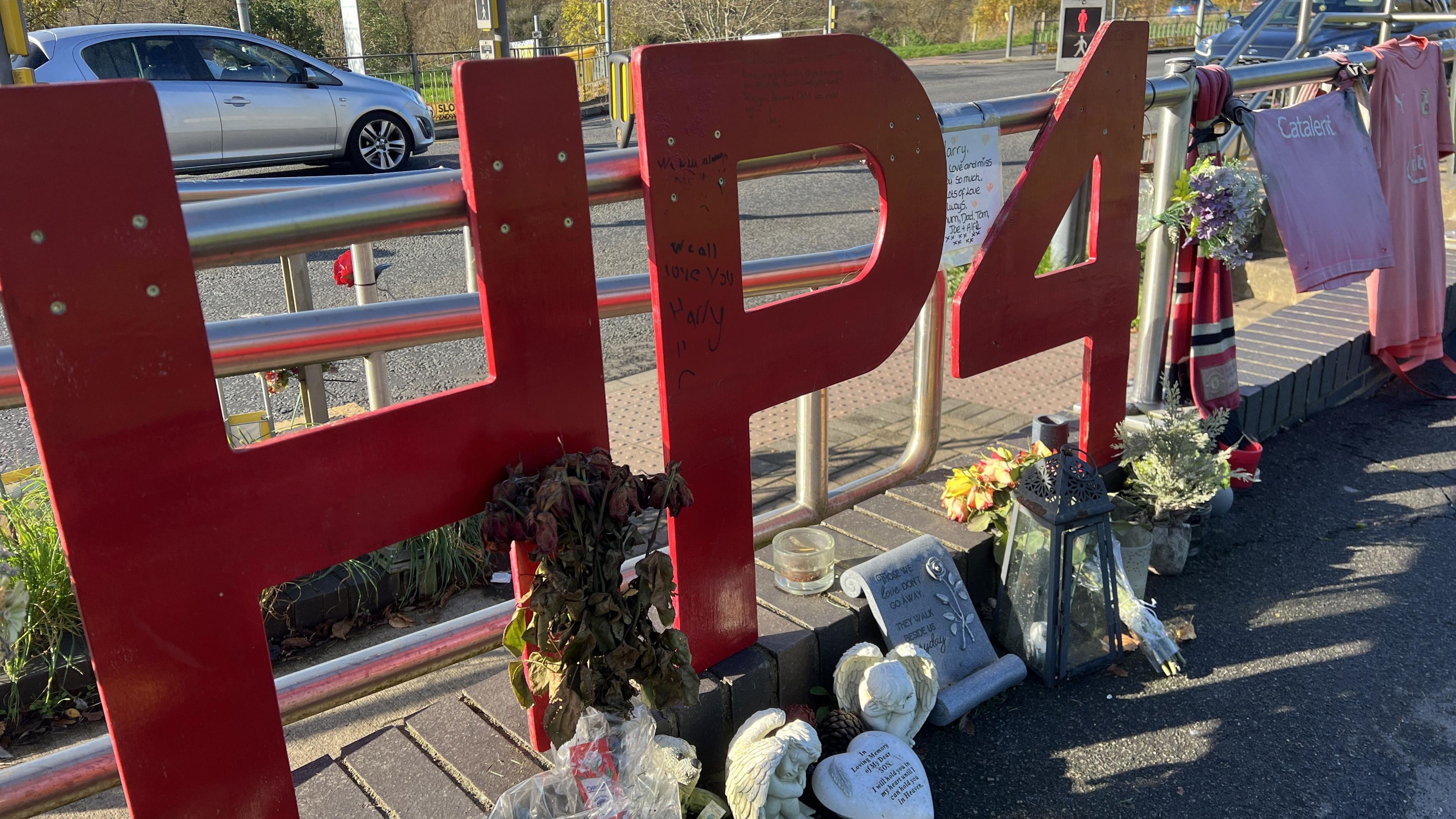 Large red "HP4" letters fixed to railings at pedestrian crossing that stand for Harry Parker and his football shirt number, alongside other football shirts, flowers, candles and other tributes that make up a memorial to the teenager on Akers Way.