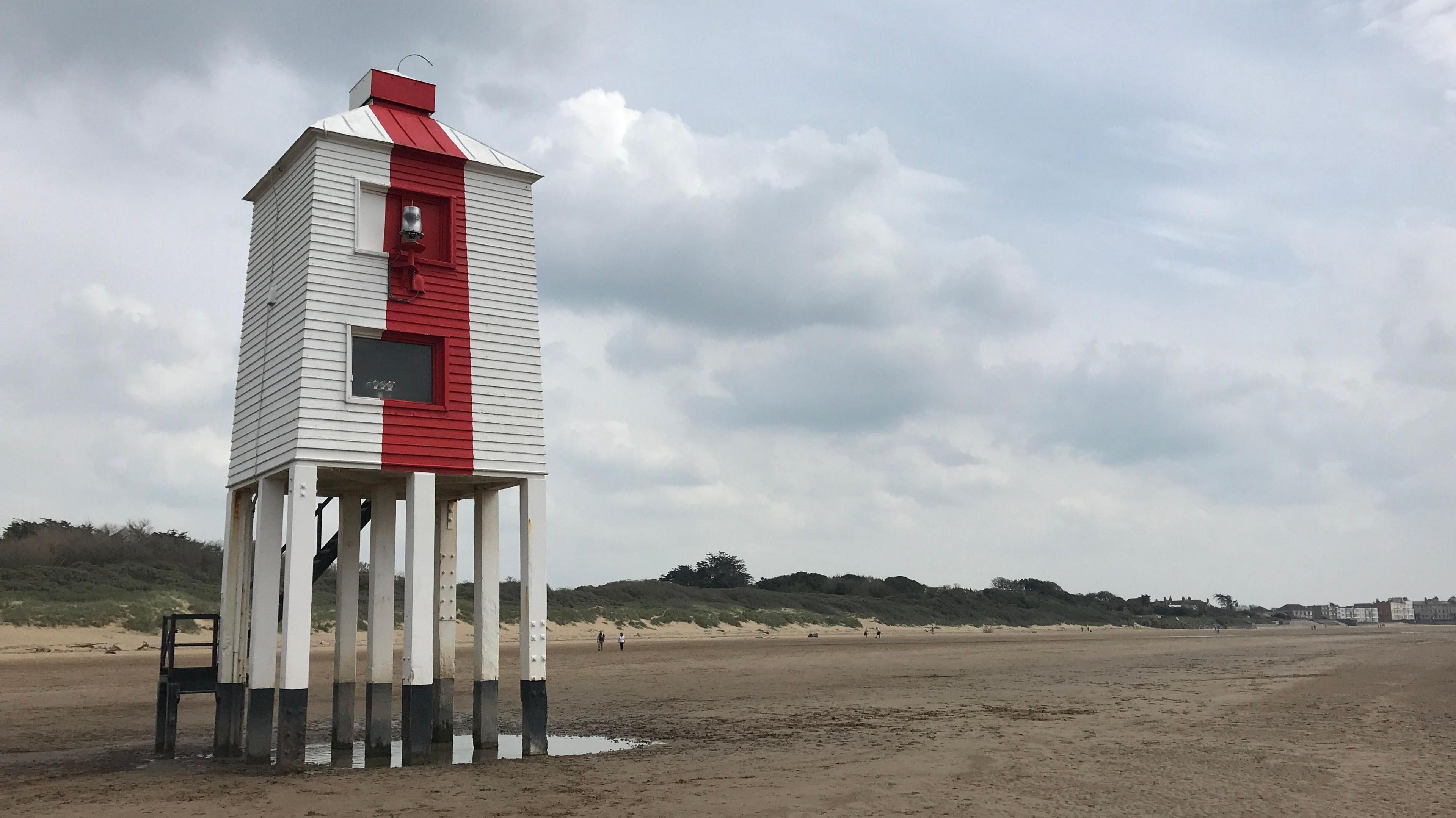 Red and white striped lighthouse on a beach. There's sand dunes in the background and a beach. It's quite a cloudy day with blue sky peeking through. 