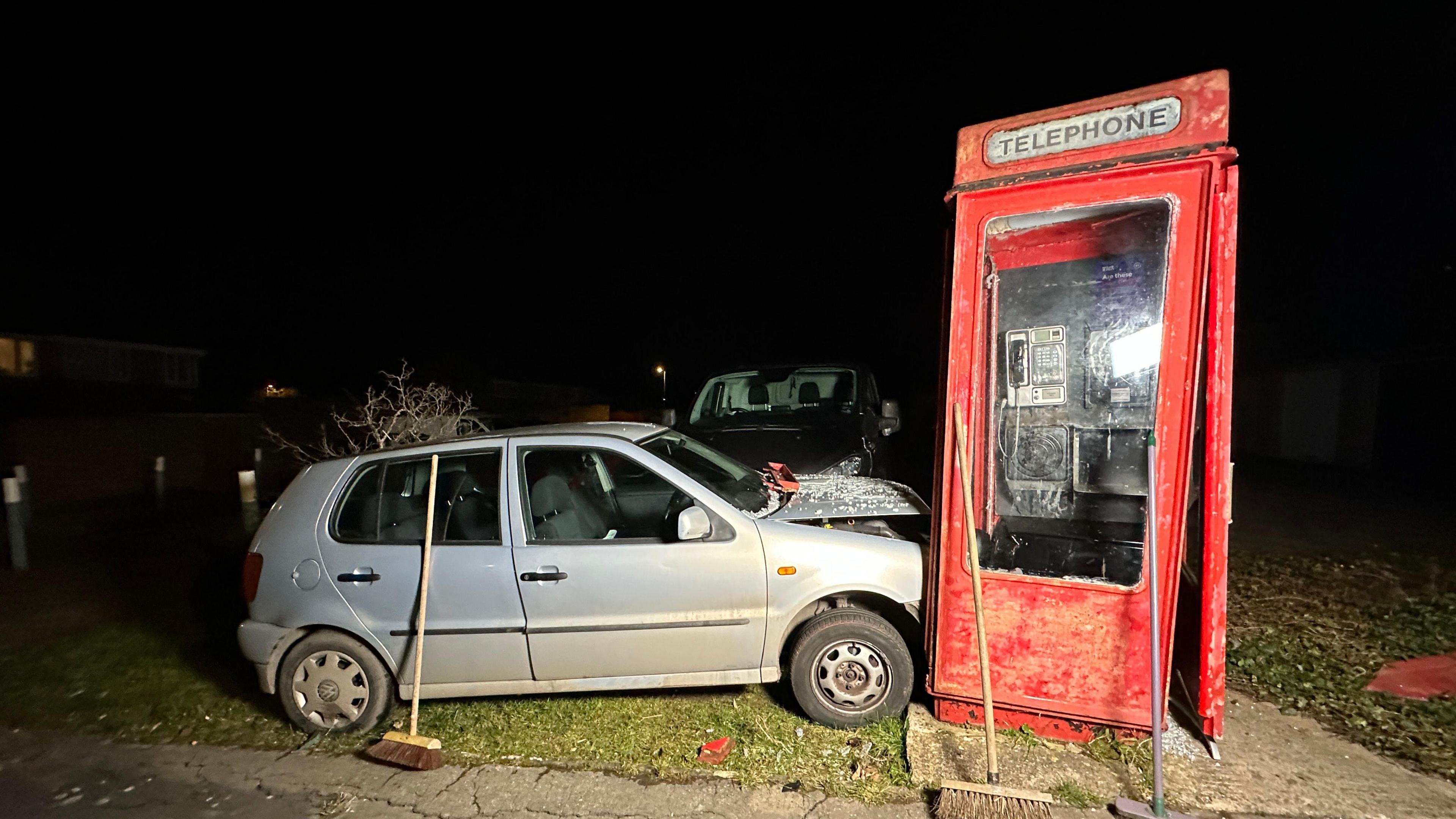 A picture at night of the red phone box, with one side coming away and leaning slightly. A silver VW polo is in the side of it, broken glass across the bonnet which popped up with the impact.