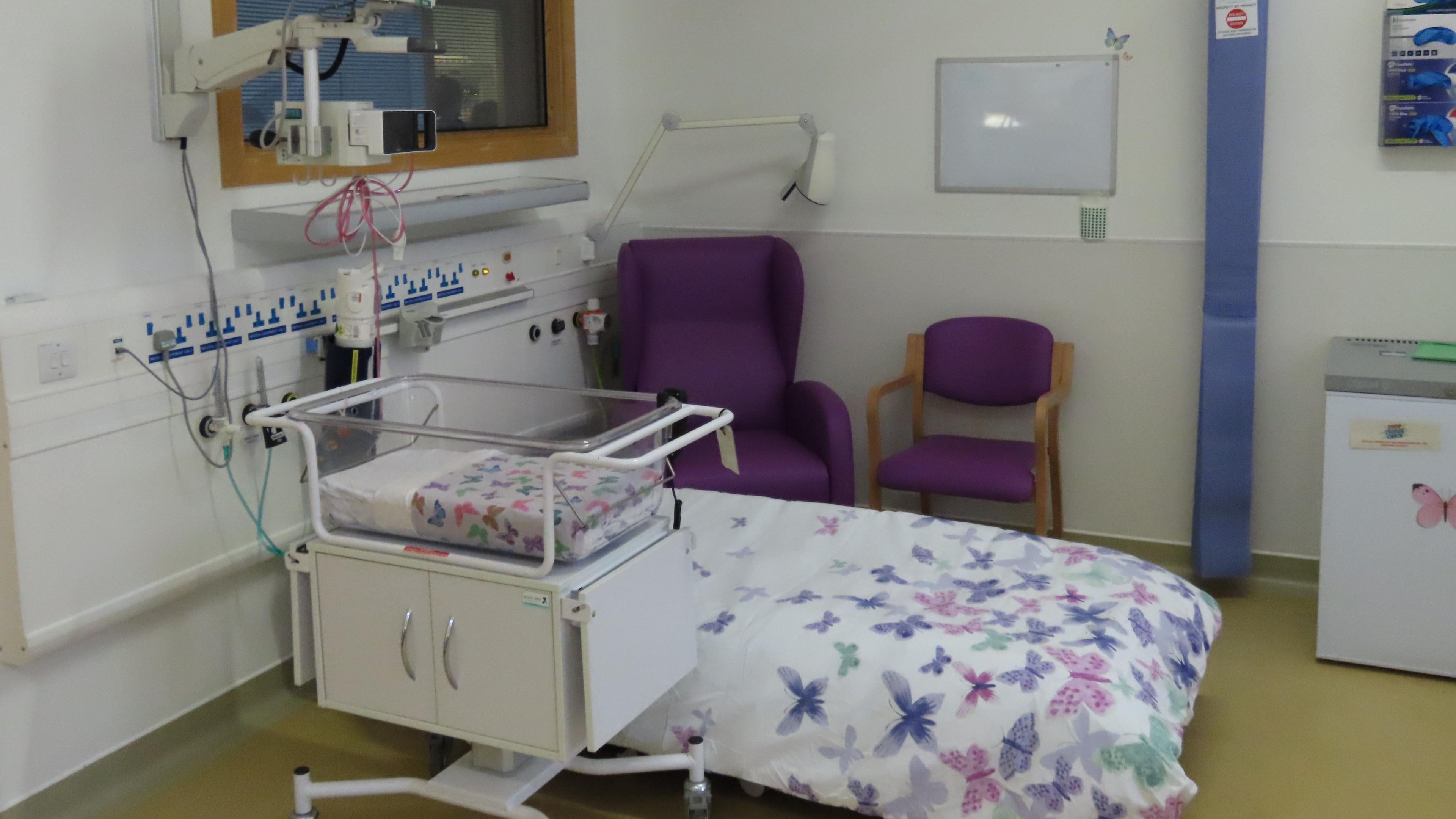 A hospital room with a bed in the centre. Next to it is a baby seethrough cot.