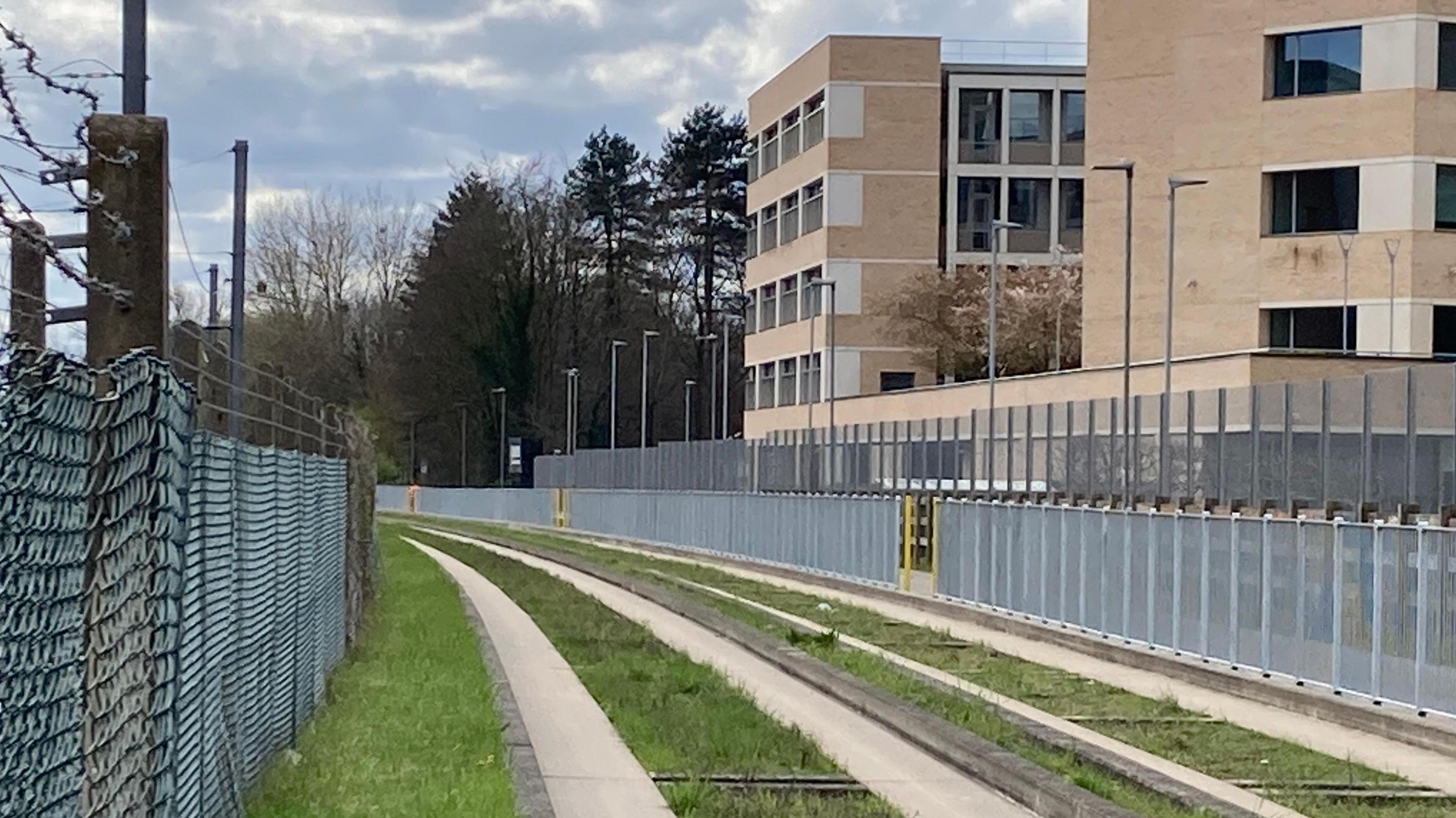 A photograph of a section of the Cambridge busway. Grass is growing on either sides of the concrete of grey concrete bordered by green grass. A line of grey metal fencing runs along both sides of the busway. It is a blue sky day but with some white clouds.