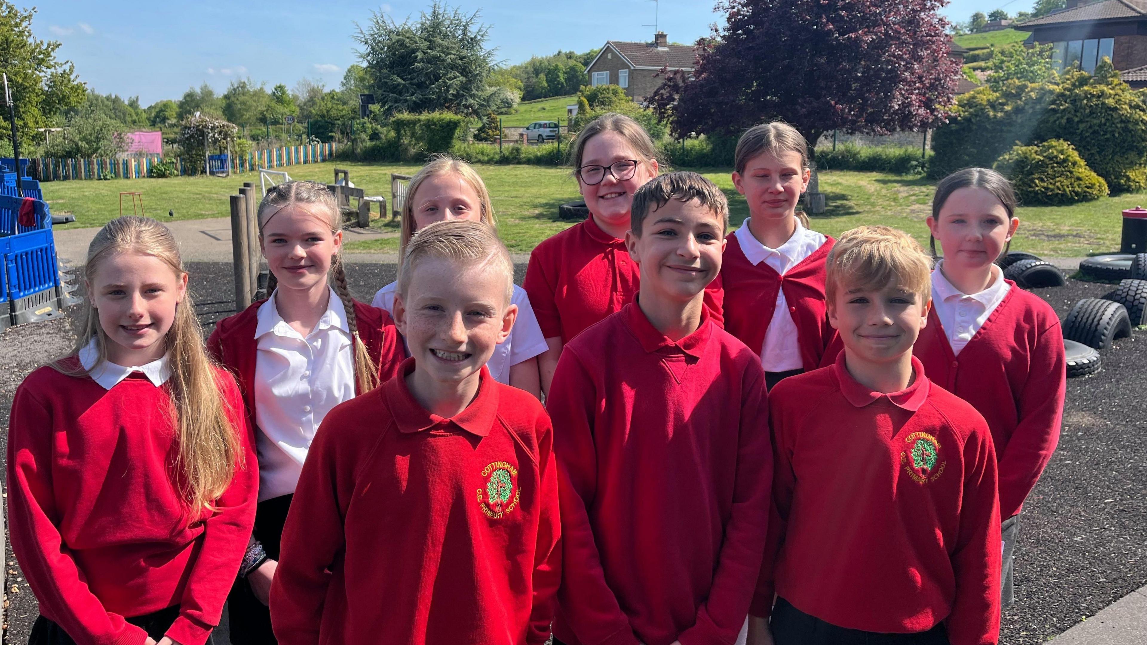 Nine children in red jumpers and white polo shirts standing on a playground with trees and grass in the background.