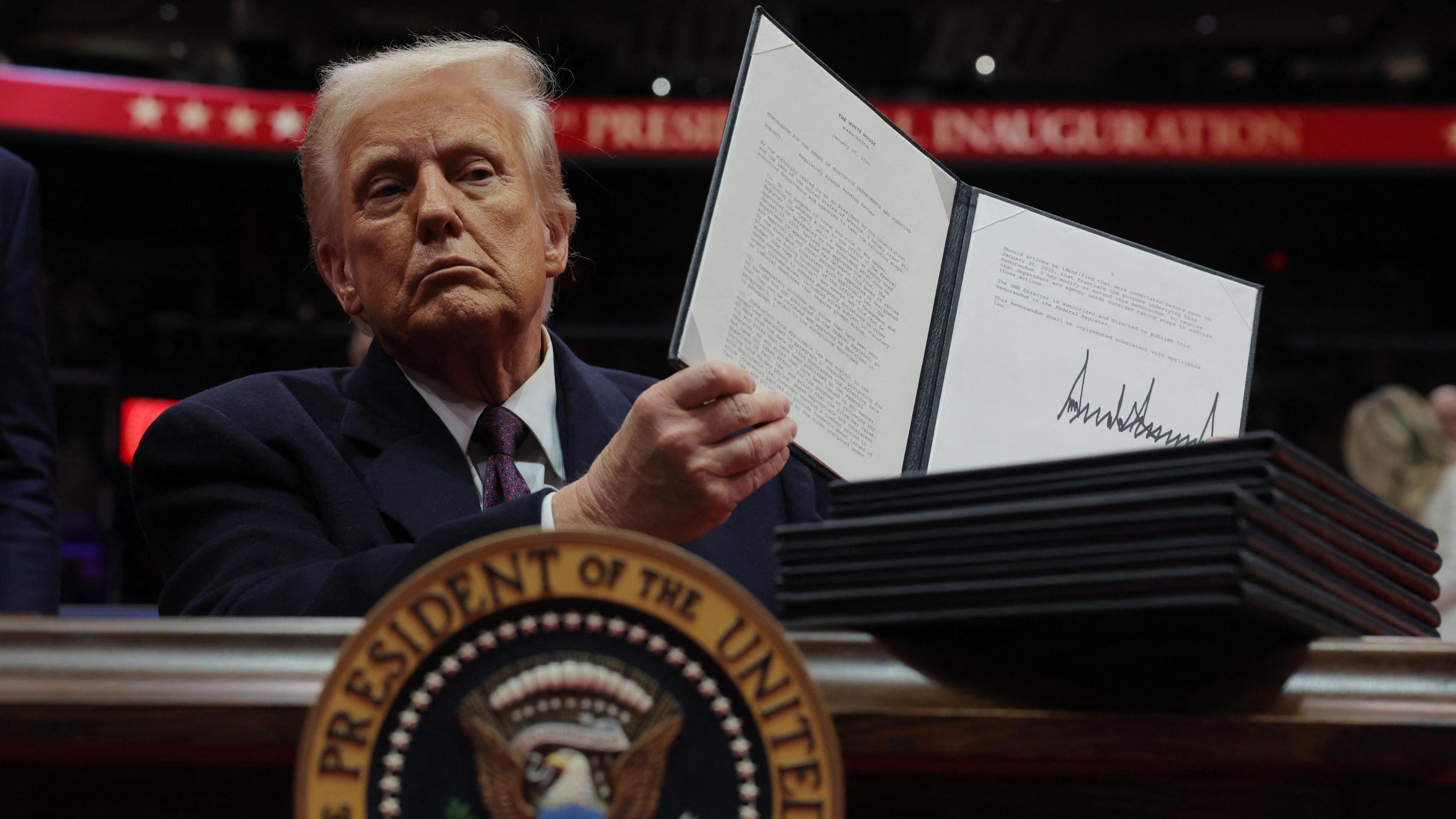 President Trump dressed in a dark suit with a dark tie and white shirt holds up a folder containing an executive order he has signed. He is sat a a table with the presidential seal in the foreground