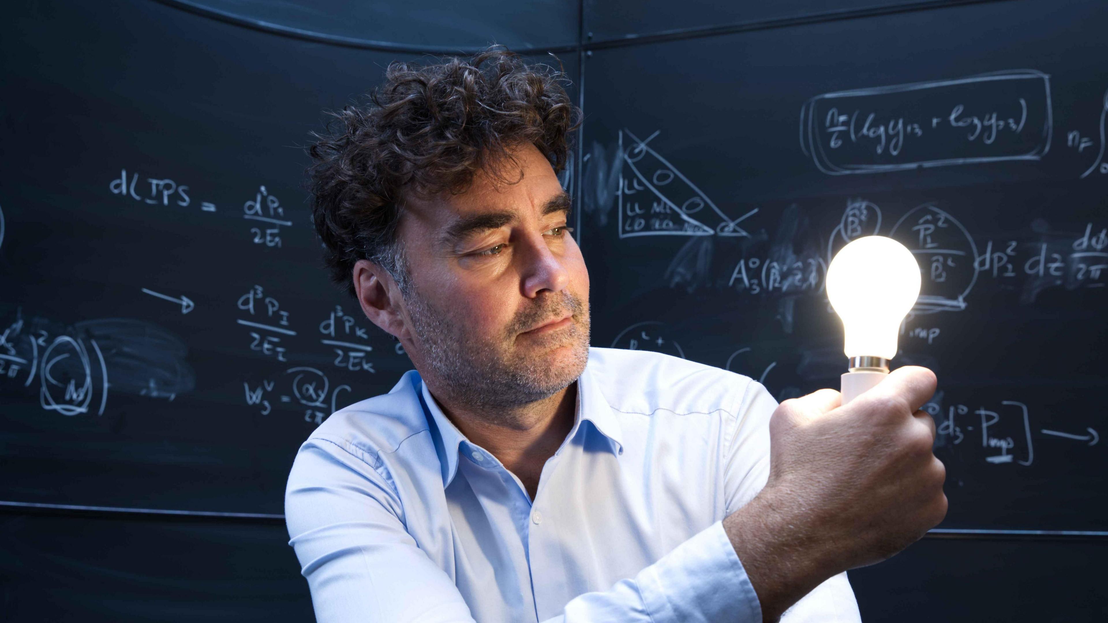 Henry Snaith is wearing a light blue shirt and has short brown hair and brown stubble. He is holding a lit up lightbulb and behind him are chalkboards with mathematical formulas written on them