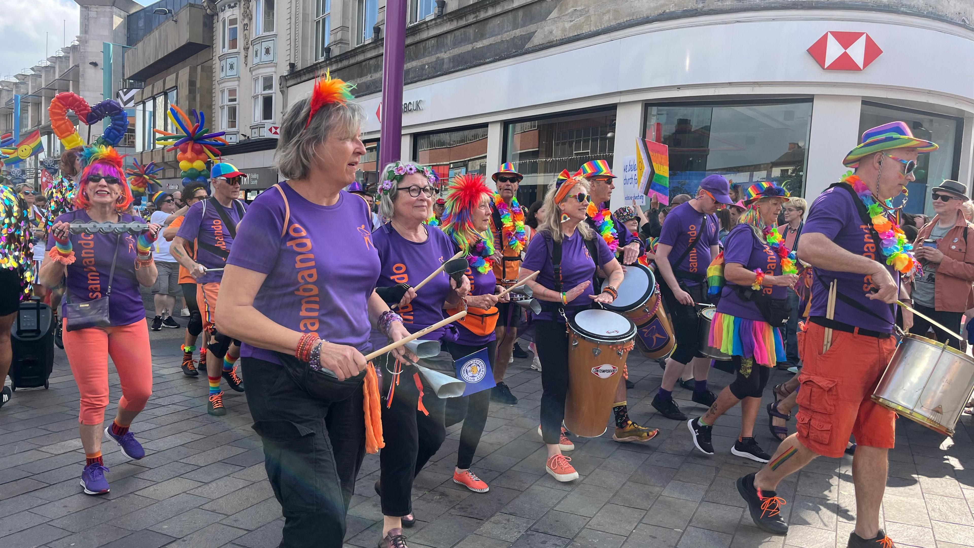 A band of drummers and percussionists wearing multicoloured accessories walking through Leicester city centre in the Pride parade