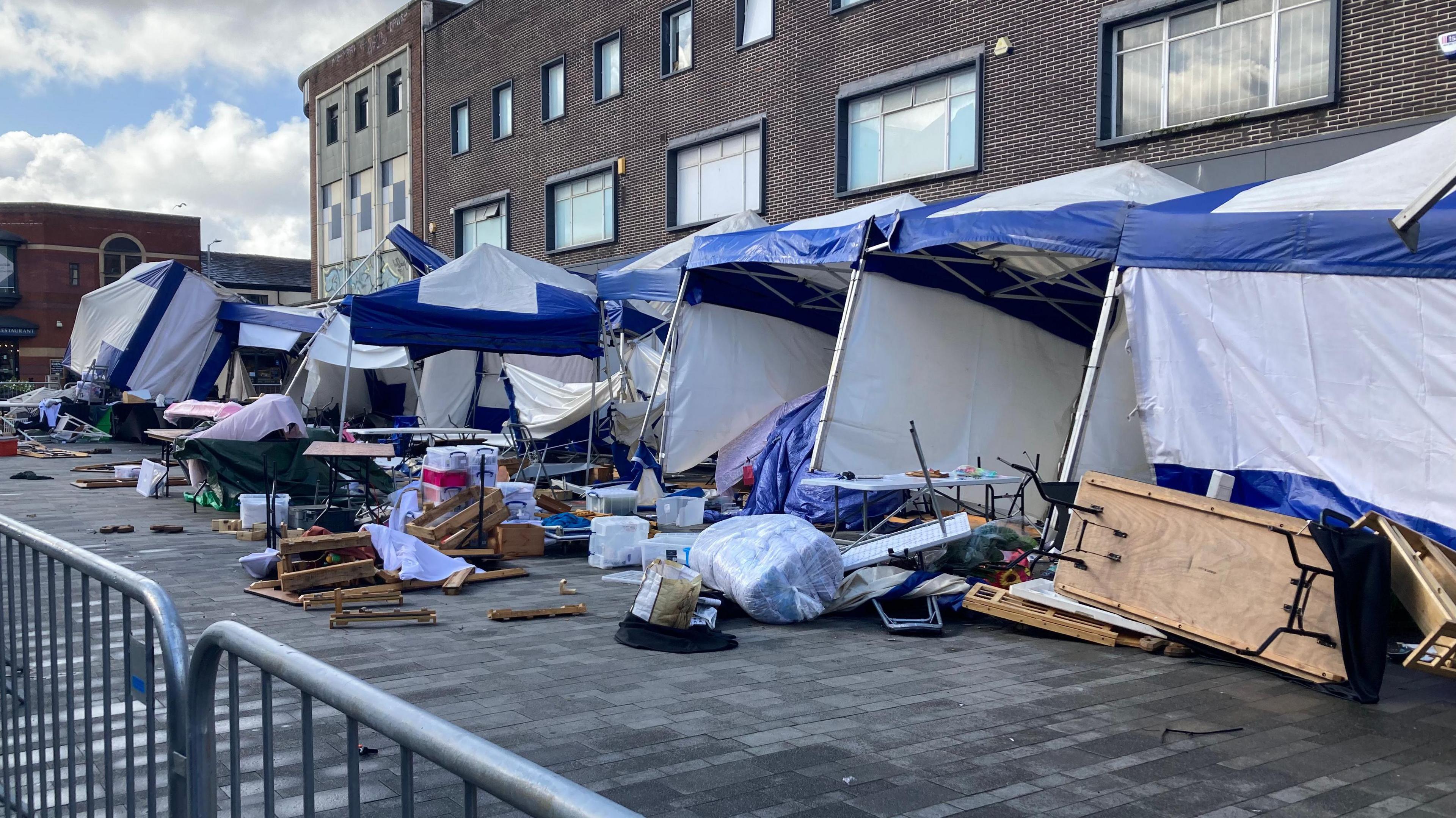 Market stalls are seen blown into shopfronts on Newport Road in Bolton, with tables upended and stock scattered across the street.