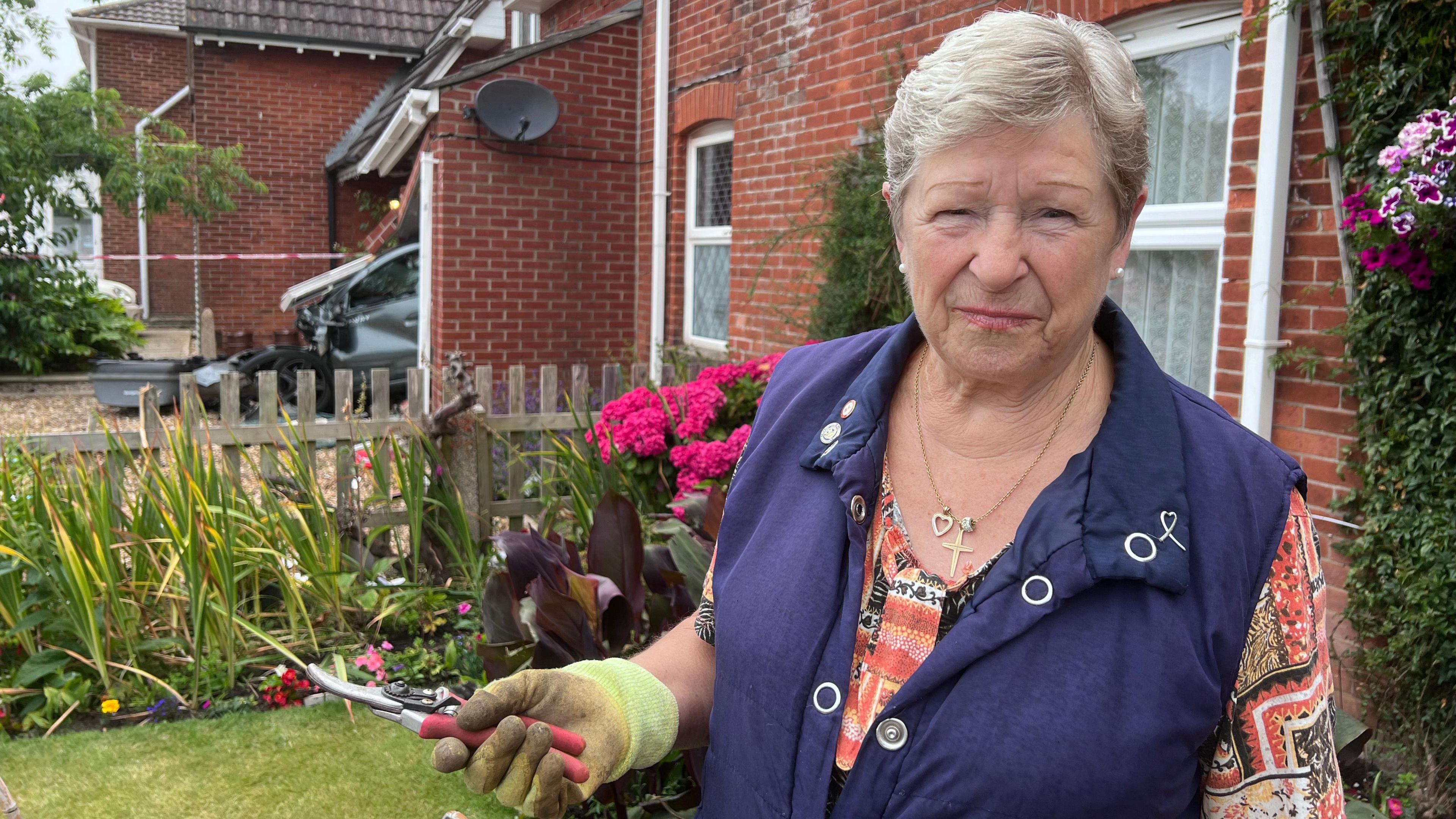 Yvonne Steele wearing a purple gilet and orange shirt, she is stood outside her home and is wearing gardening gloves and holding secateurs