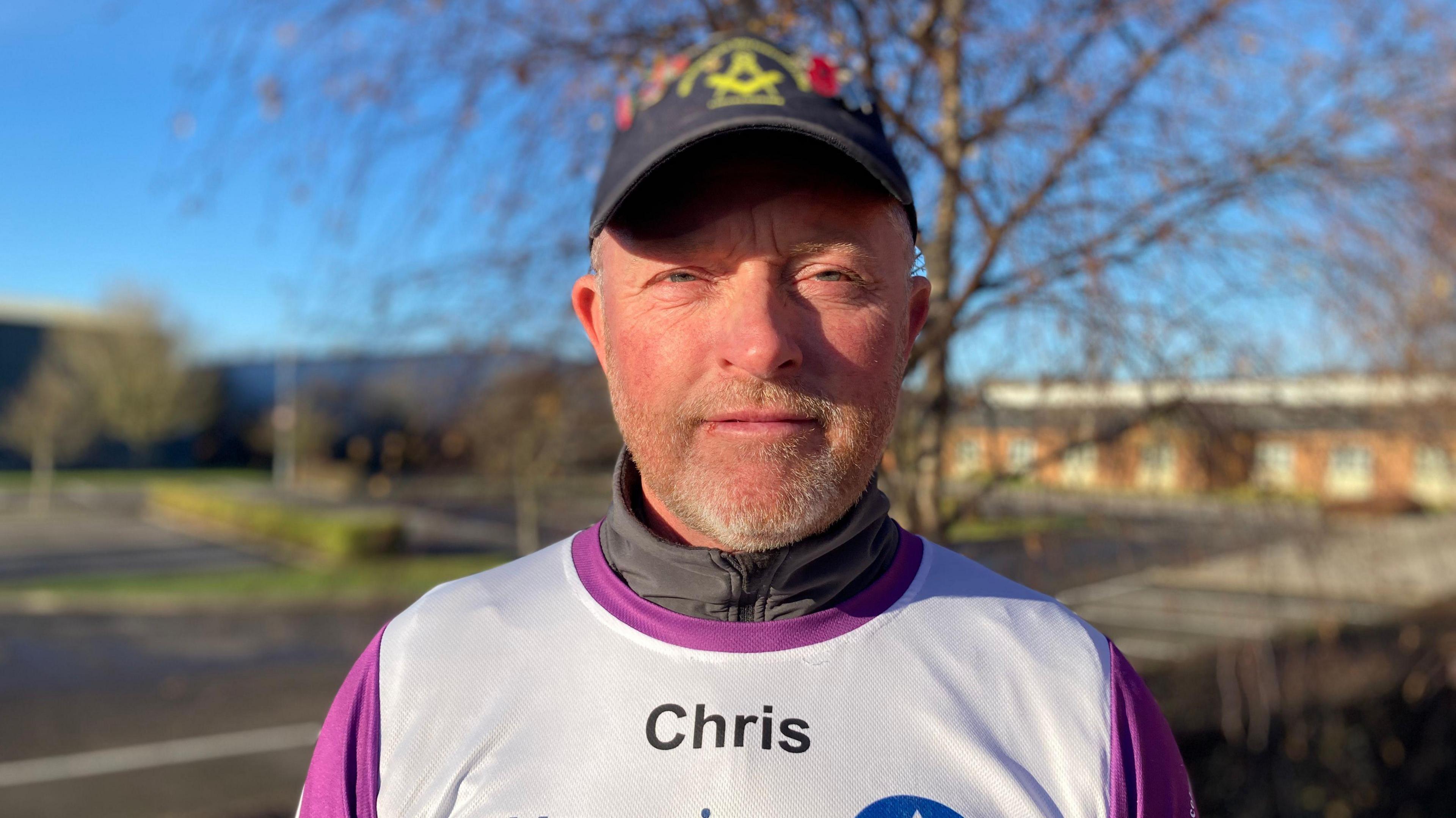 Chris smiles facing the camera in a jersey with his name on it.  He wears a cap on his head and is sporting stubble on his chin.