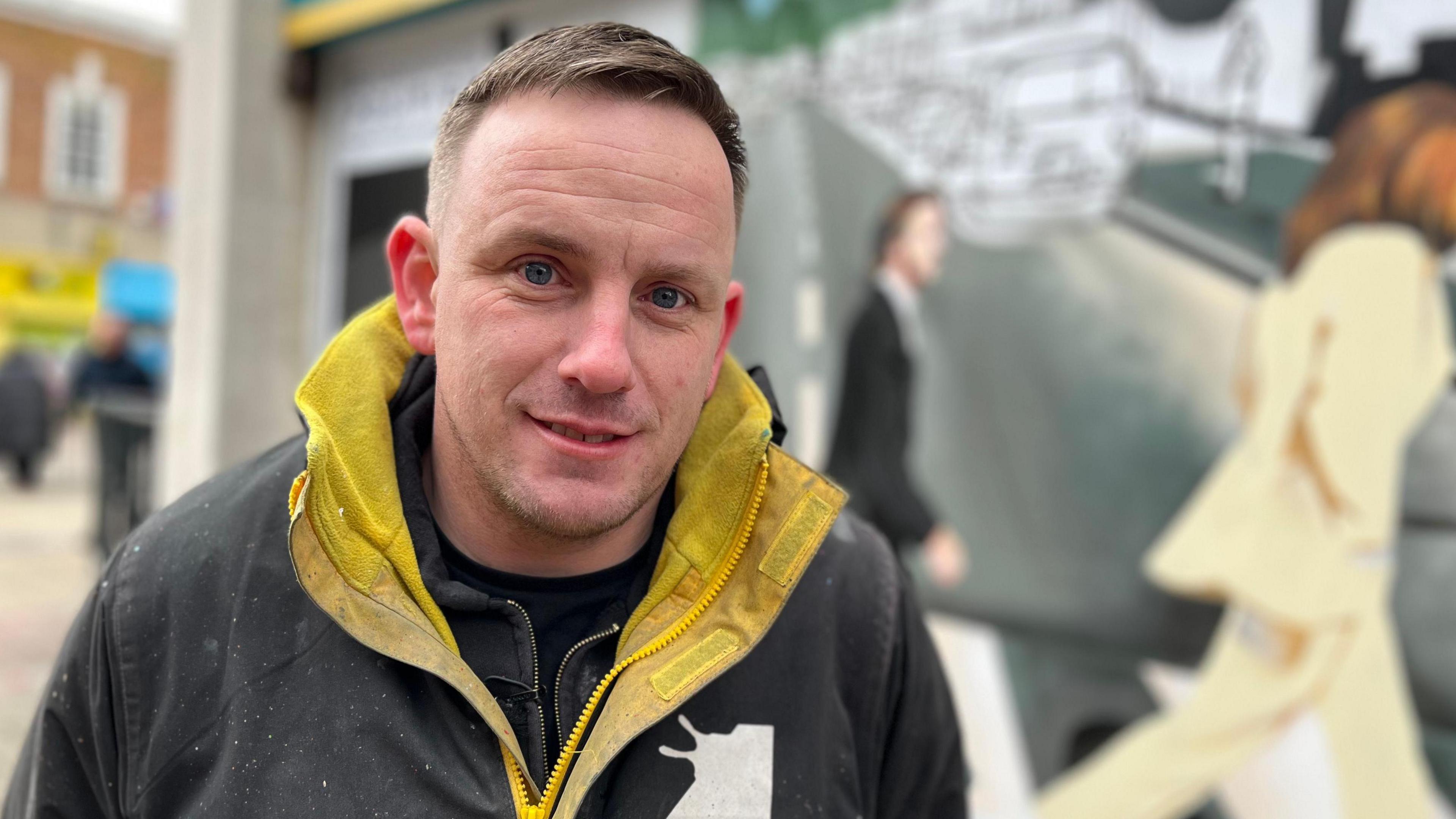 Nathan Murdoch, a man who is looking at the camera and smiling. He is wearing a yellow and black jacket and is stood in front of a mural he has been creating. 