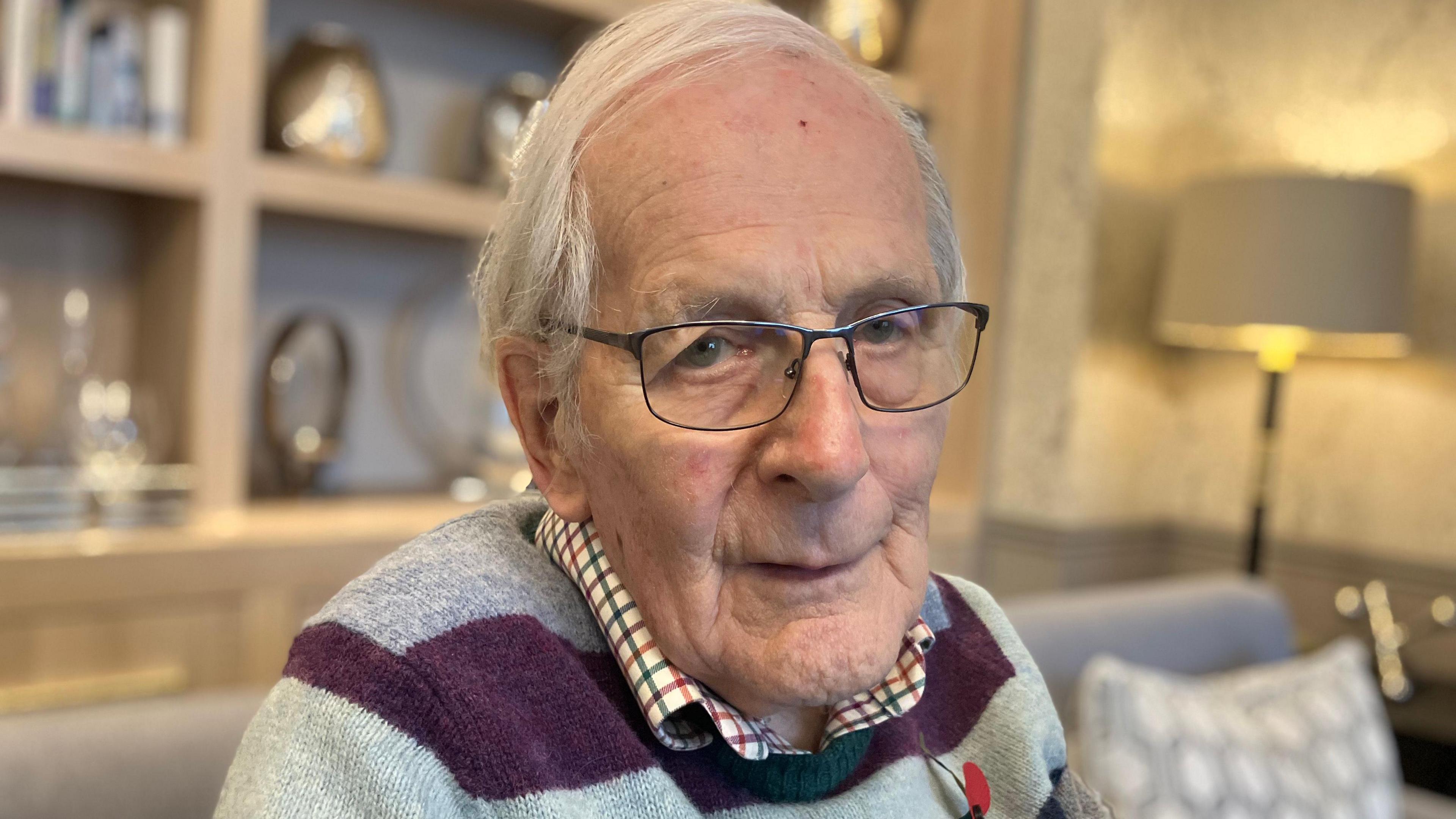 A head and shoulders photo of a man with grey hair and glasses and wearing a purple and grey stripy jumper. There's a lamp and a set of shelves in soft focus behind him.