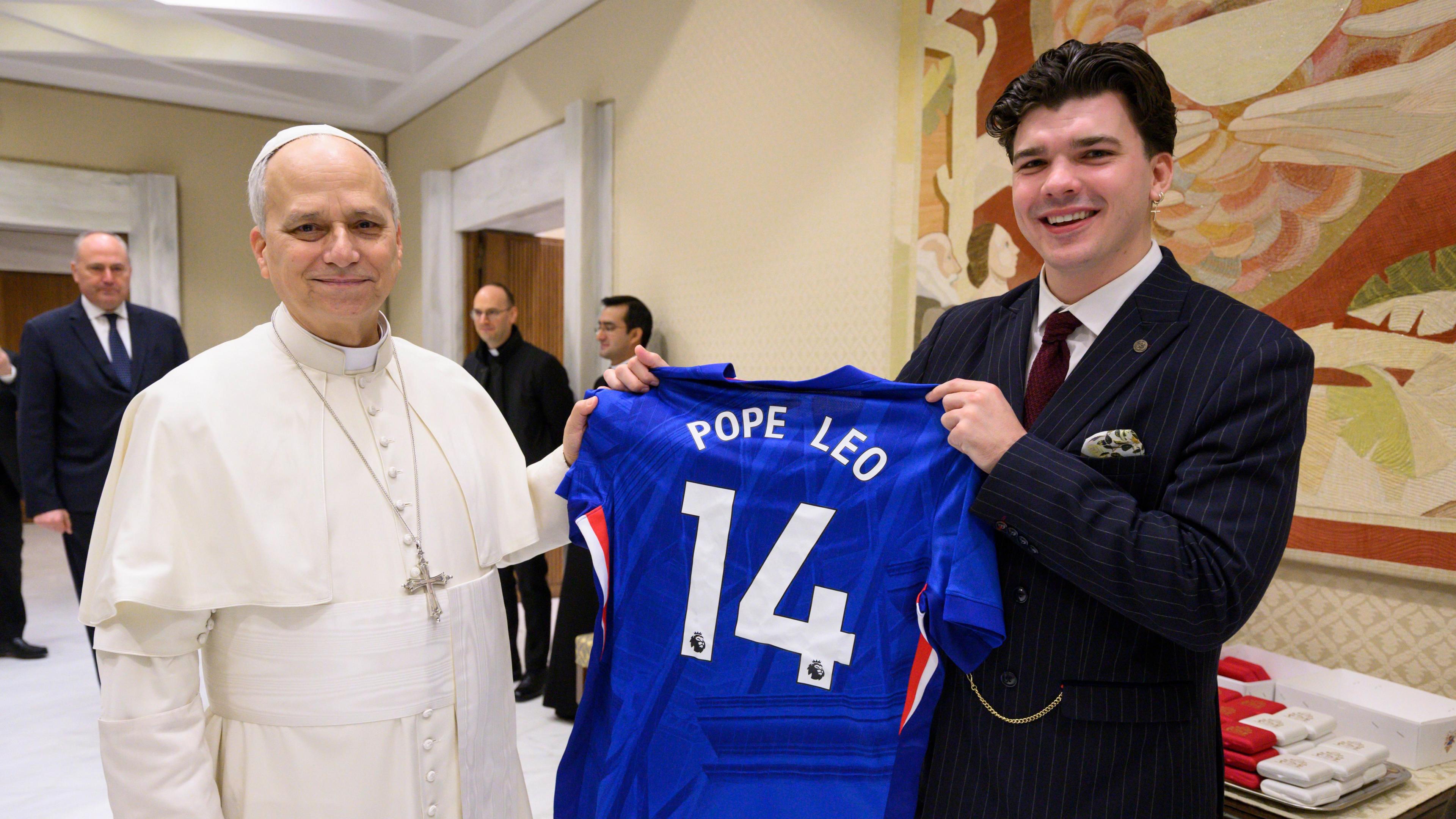 The Pope dressed in white stands next to Harry Clark in a black suit, they are both holding part of a Chelsea F.C. shirt that has Pope Leo 14 written on it.