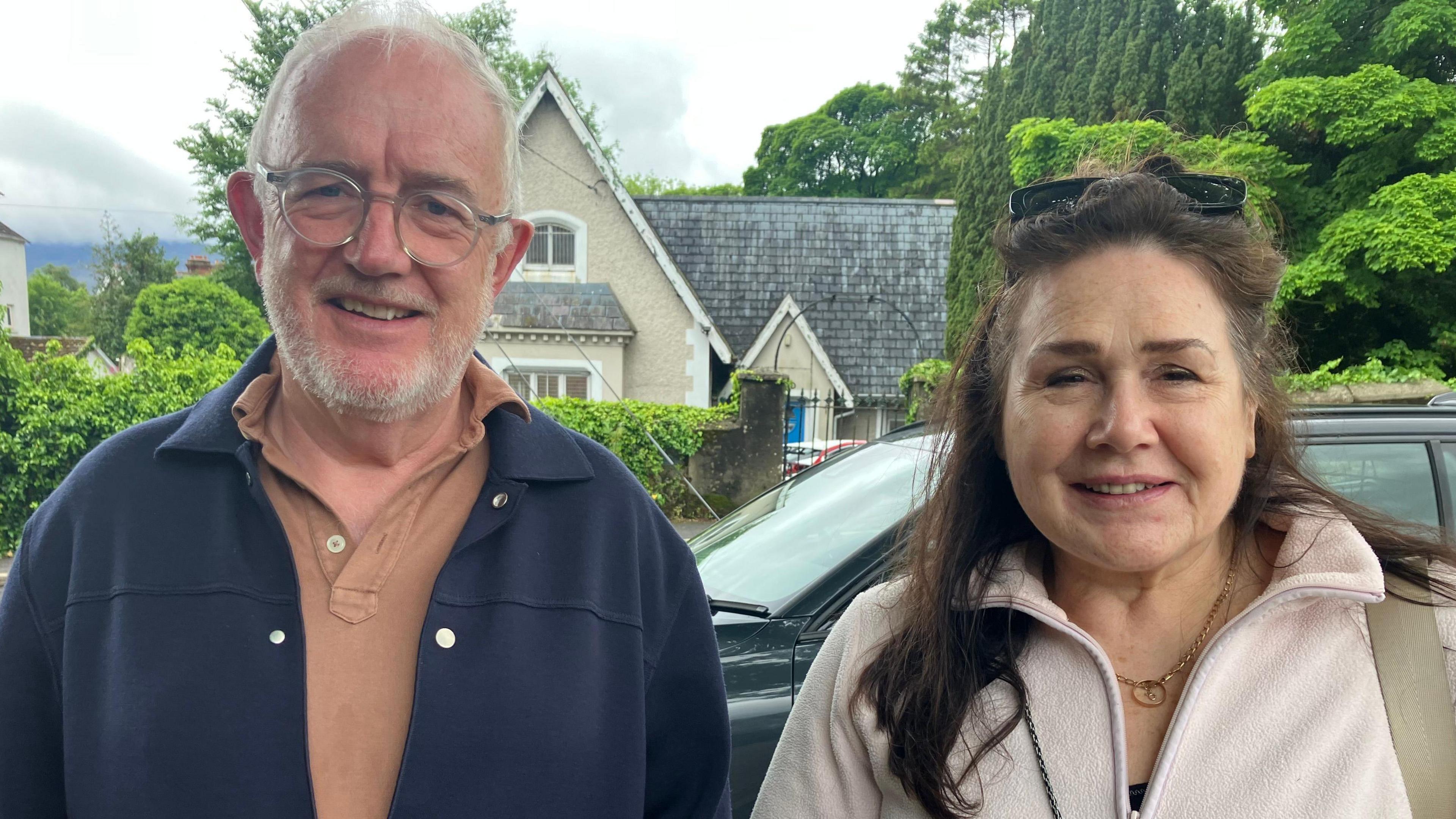 Pat Fitzgibbon and his wife Aisling Donohoe looking to the camera in Rostrevor. Pat has short, white hair and a beard and is wearing glasses, a navy jacket and a brown polo shirt. Aisling has long brown hair and is wearing a pink fleece and a gold necklace.