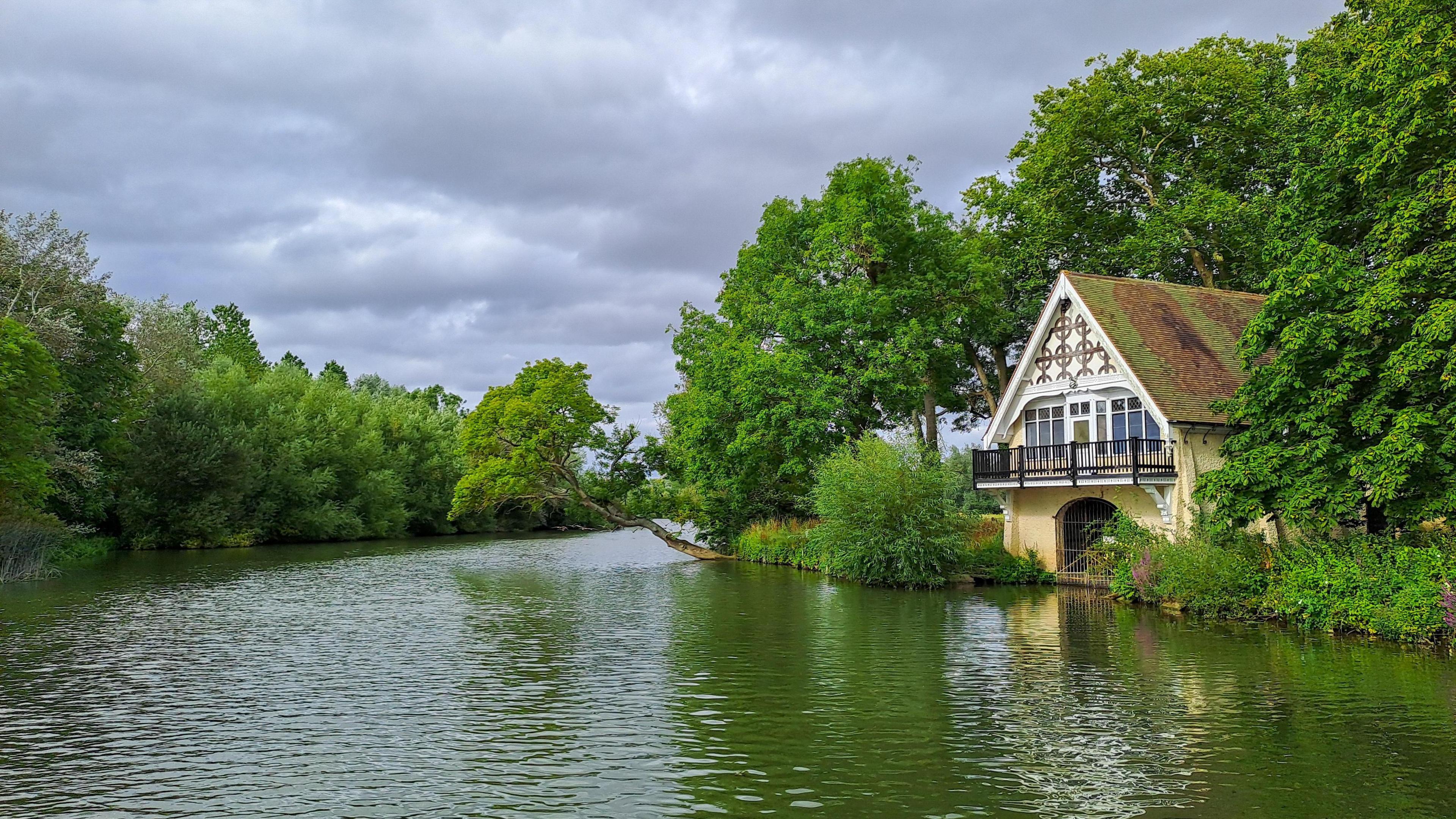 A cream and white boat house with a pitched roof pokes out amongst trees from the right of the image. It has a balcony and a gated entrance at water level. The river reflects the green of the trees either side of it and the cloudy skies above.