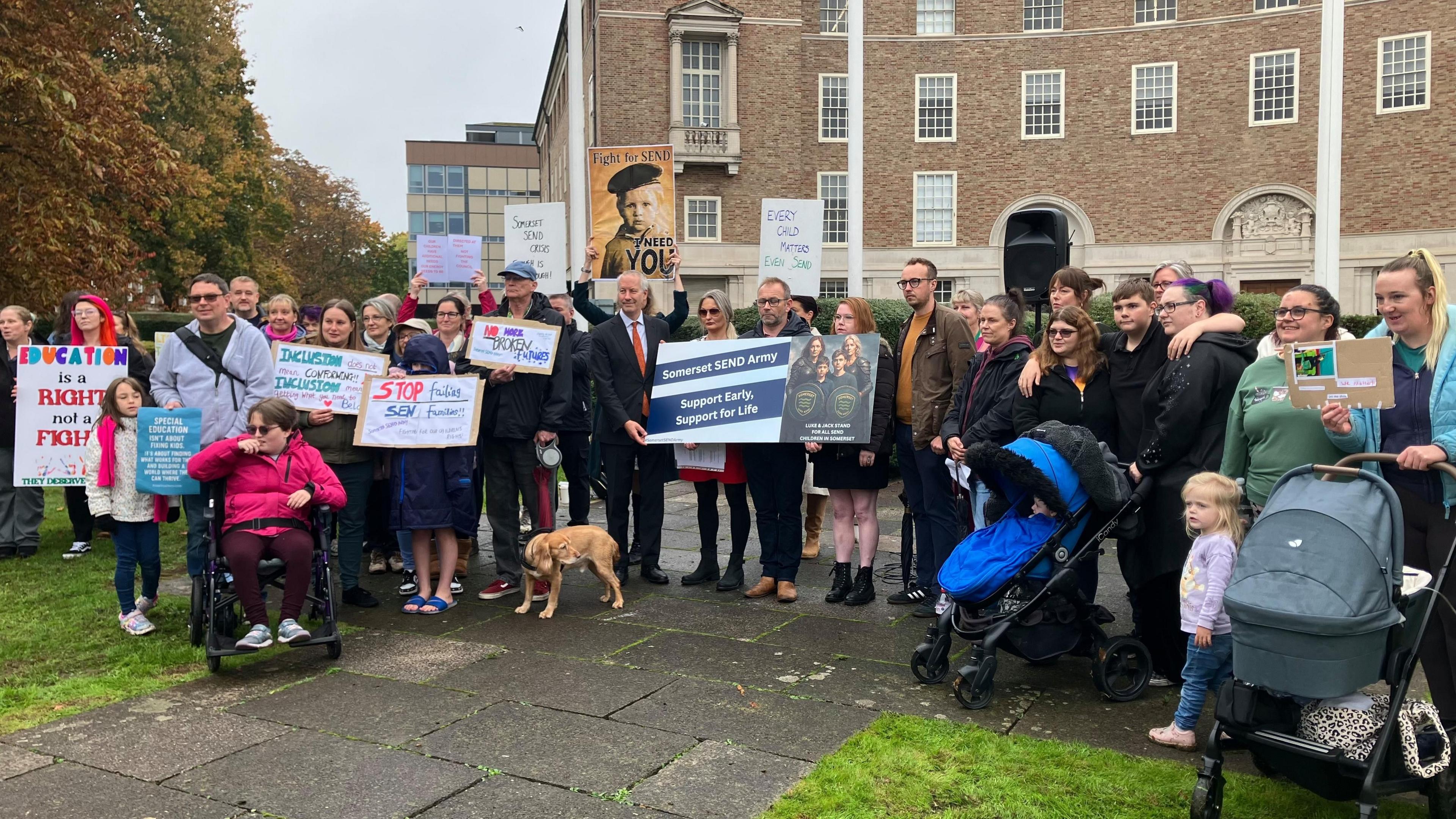 A group of people including children in pushchairs, wheelchairs and a dog on a lead, standing on grass outside a municipal building holding placards 
