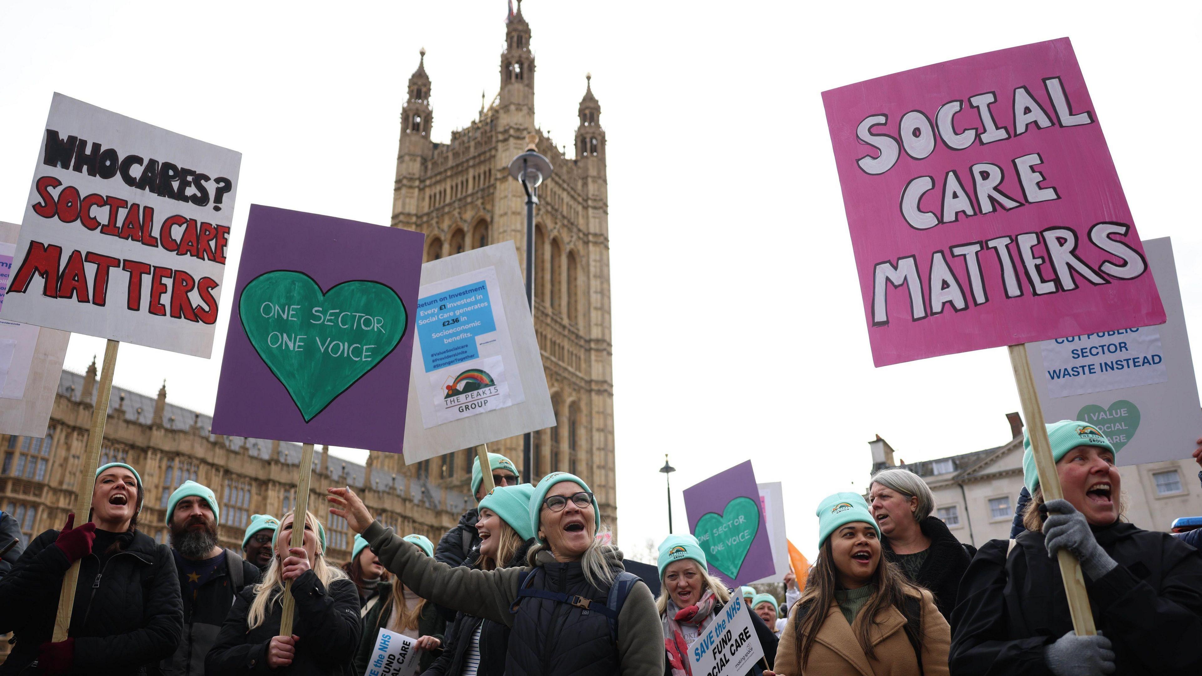 A group of people holding placards and wearing matching turquoise woolly hats outside of the House of Parliament